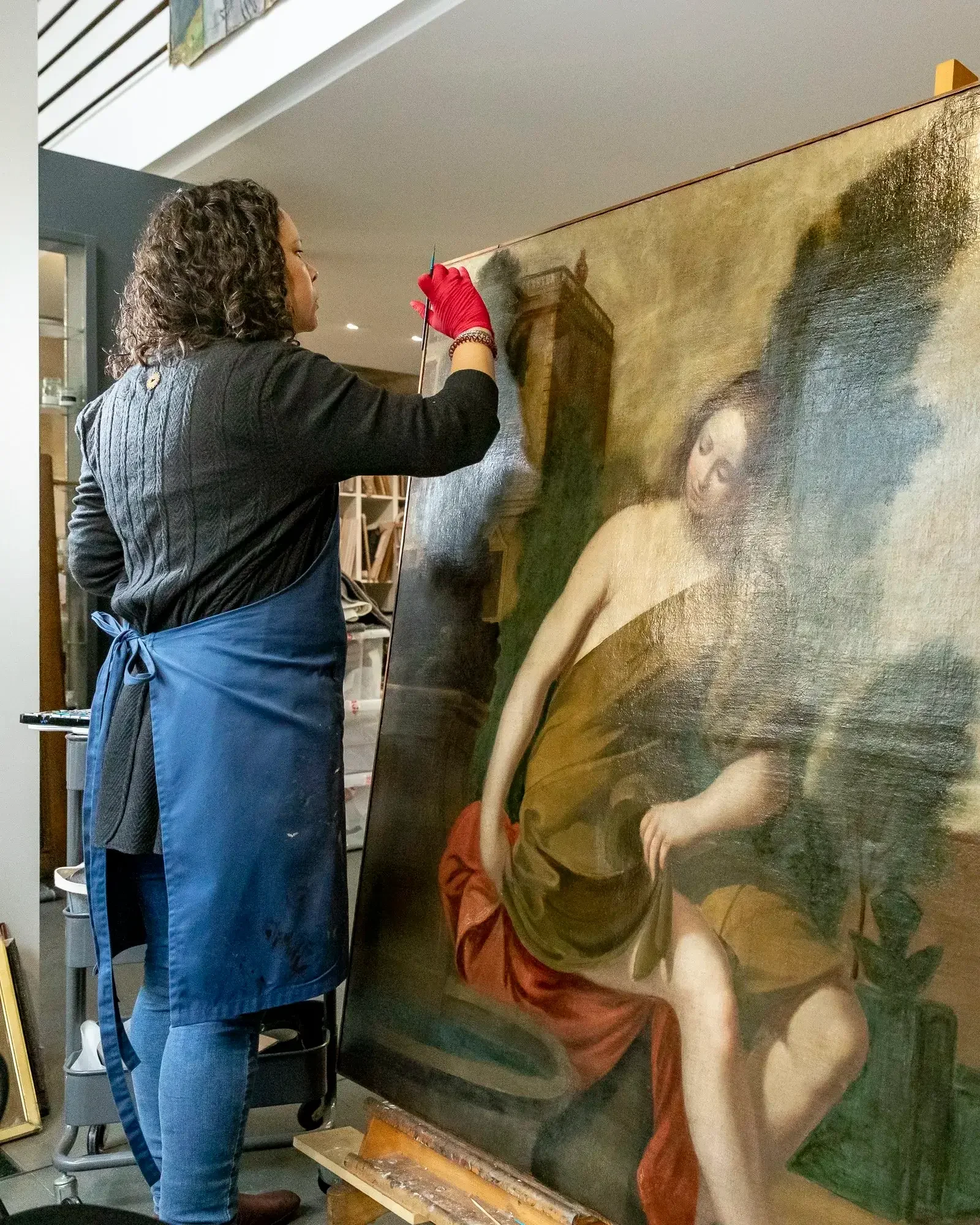 Une restauratrice travaille sur une grande peinture représentant une femme assise, portant un vêtement beige et rouge, dans un atelier.