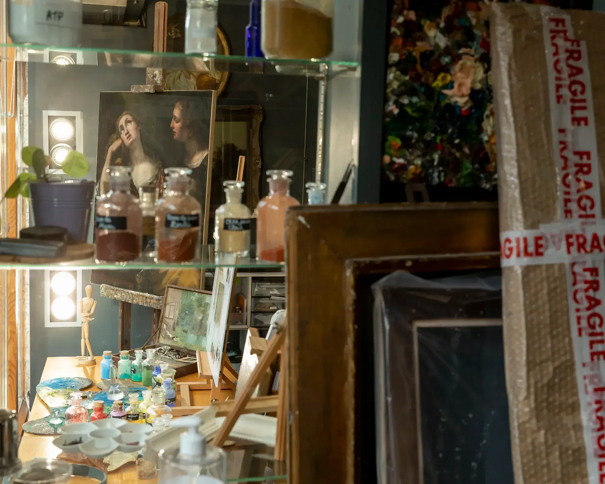 Une salle d'artiste avec des peintures, des pinceaux, des pots de peinture, et une sculpture. On aperçoit également un tableau avec deux femmes lors de la peinture.