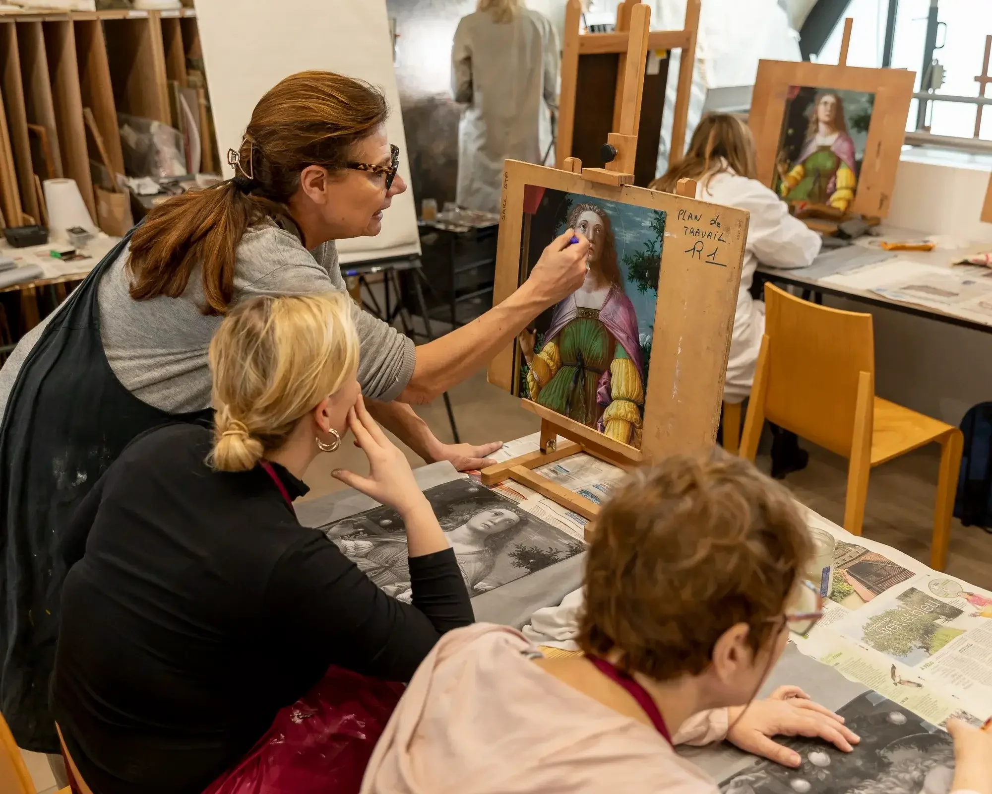 Une femme enseignant une leçon de peinture à un groupe de personnes dans un studio d'art, en train de montrer un tableau sauvegardé. Des élèves regardent attentivement, certains prenant des notes ou observant le tableau.