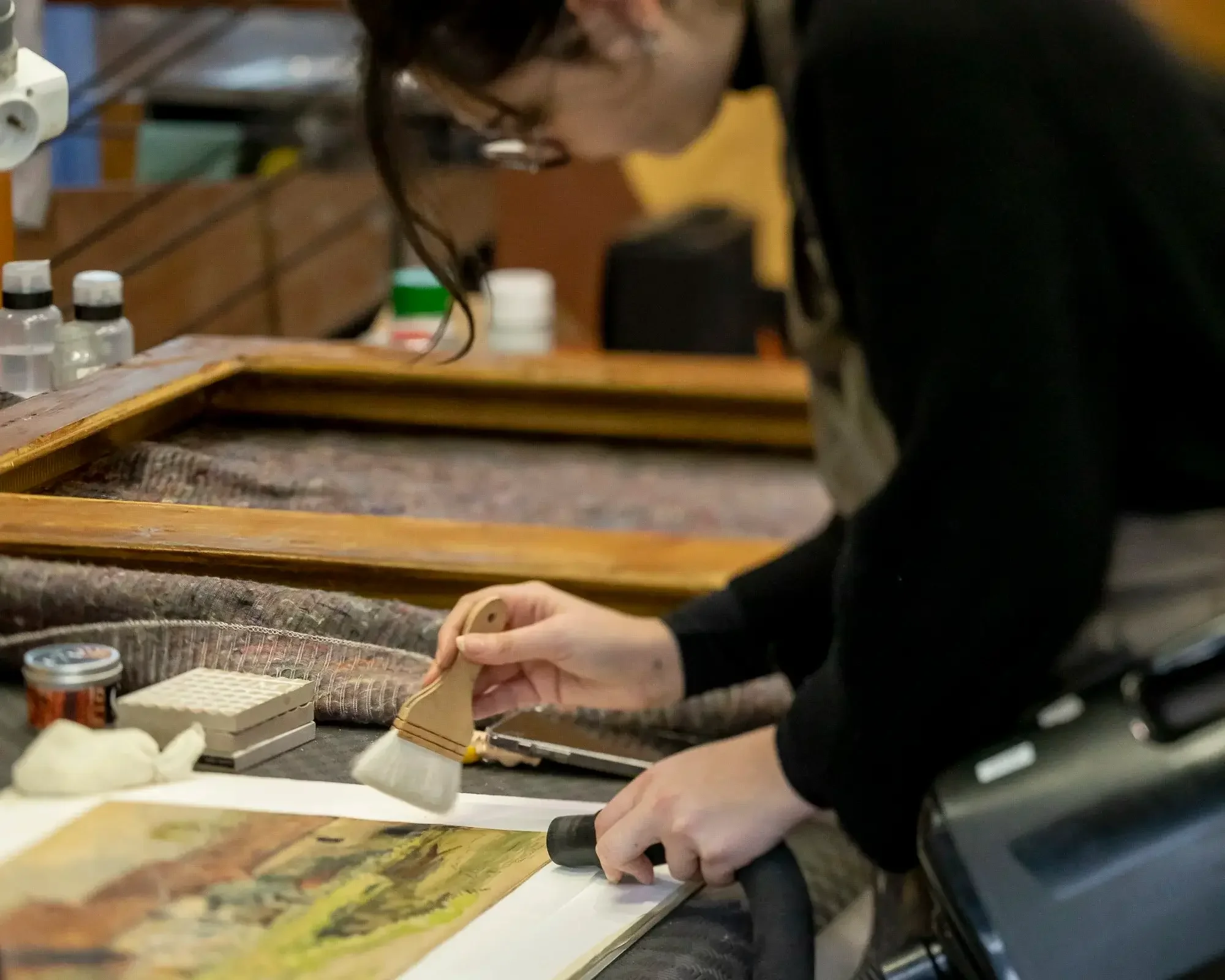 Une personne nettoyant une peinture ou une photographie ancienne sur une table de travail, avec divers outils et matériaux autour.