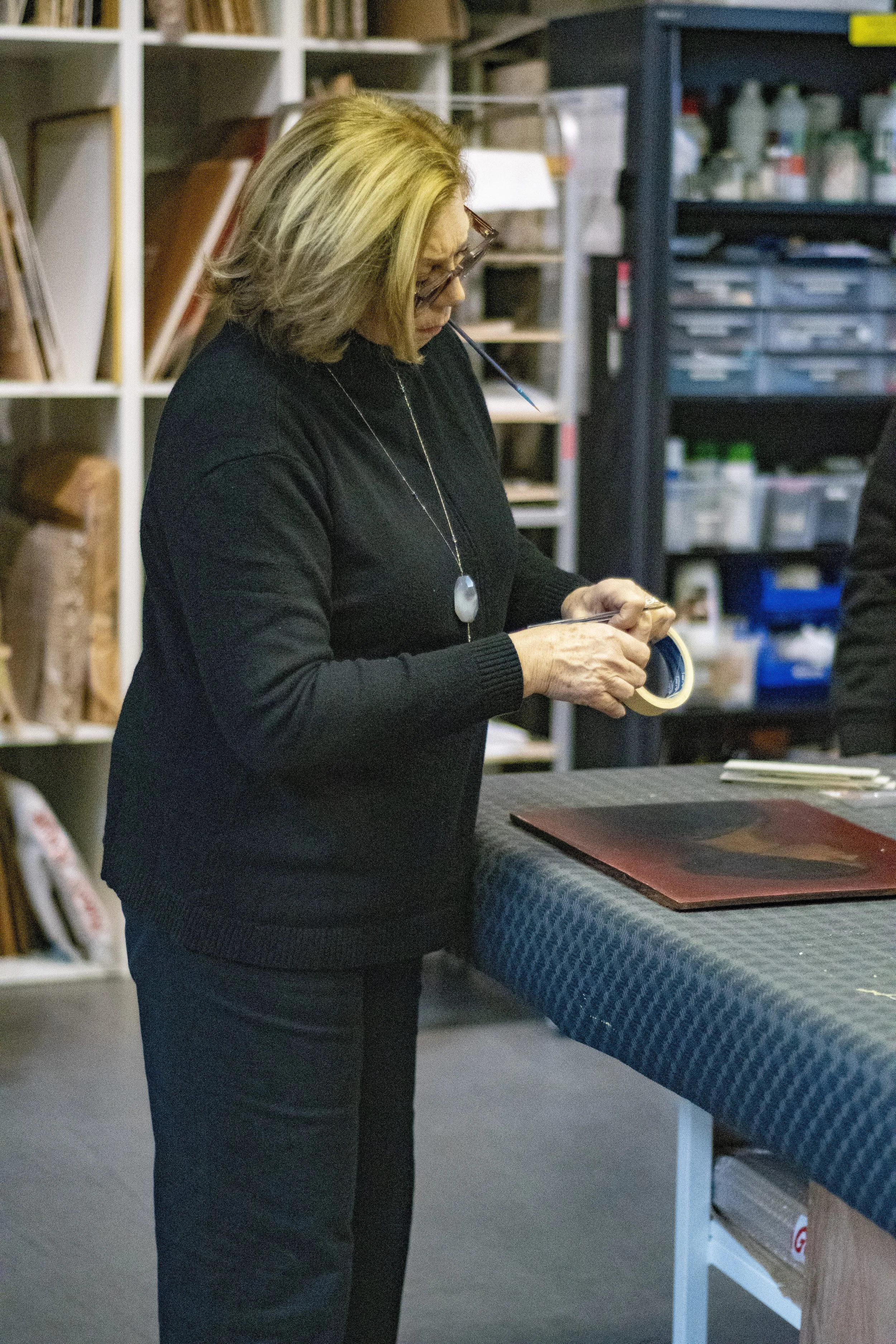 Femme blonde portant un pull noir, examinant du ruban adhésif dans un atelier avec étagères contenant des livres et des fournitures diverses.