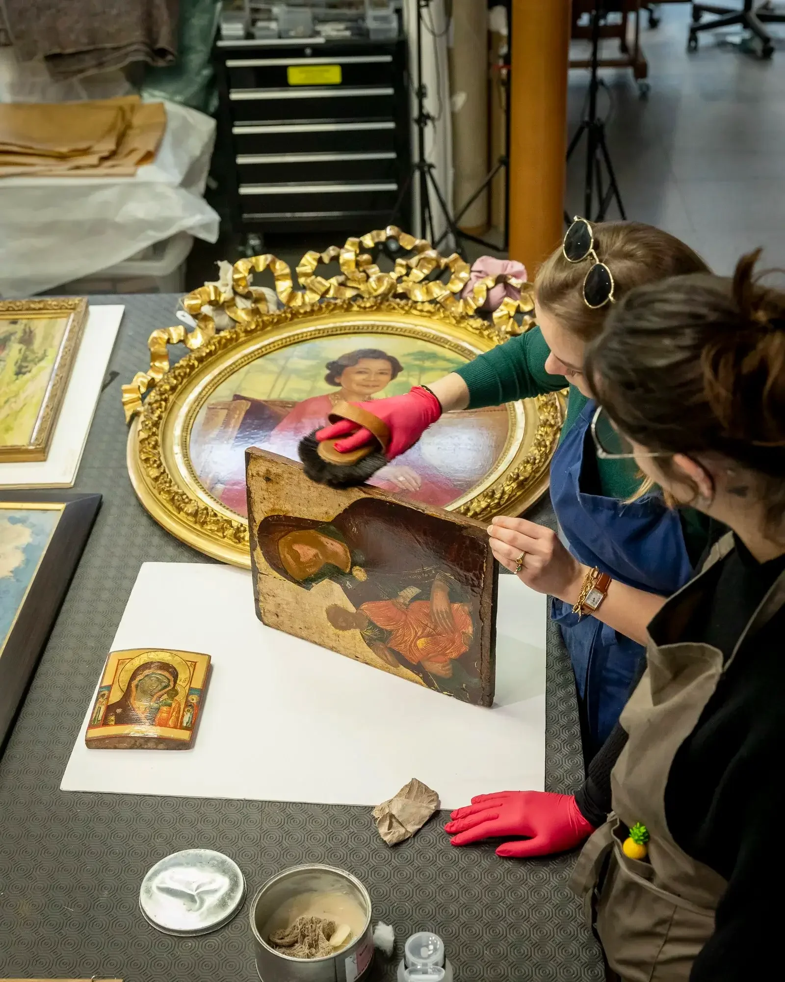 Deux personnes restaurent une peinture ancienne dans un atelier d'art. Elles portent des gants rouges pour manipuler la peinture, une d'elles utilise une brosse pour nettoyer le cadre doré de la peinture.