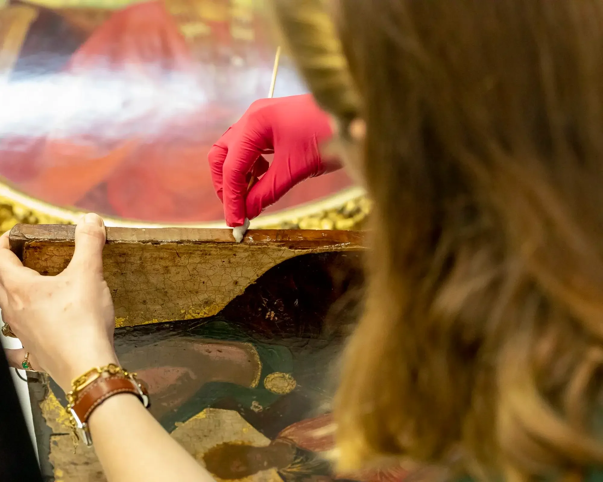 Une personne en train de restaurer une peinture ancienne, utilisant un pinceau ou un outil précis, avec un gant rose. La scène est vue de derrière, avec des cheveux bruns visibles en premier plan.