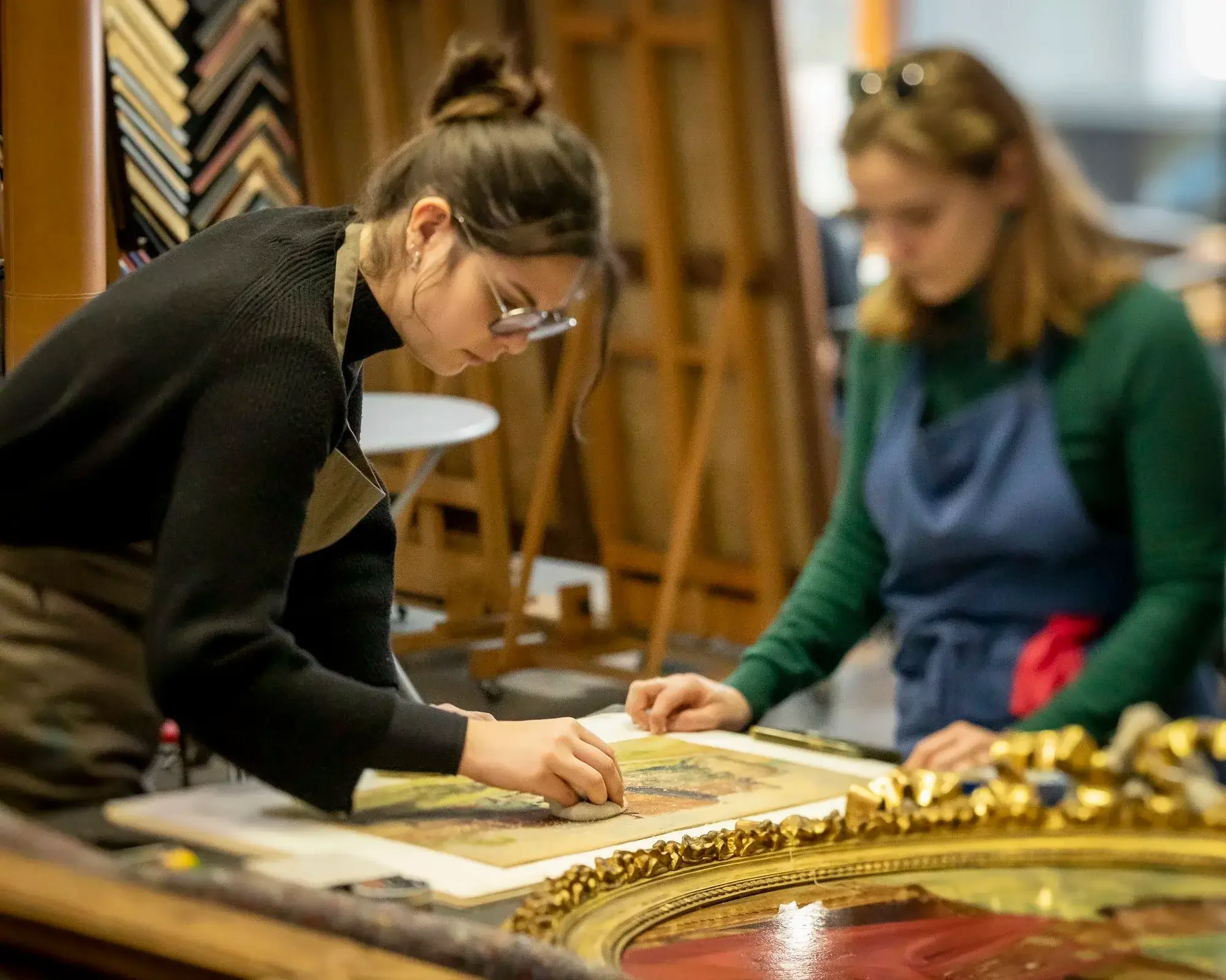 Deux femmes travaillent sur une restauration d'œuvre d'art dans un atelier, portant des lunettes de protection et des tabliers.