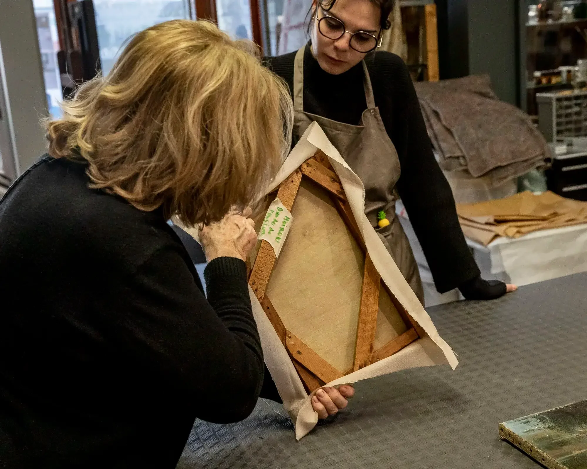 Deux femmes examinent un cadre retourné sur une table, dans un atelier.