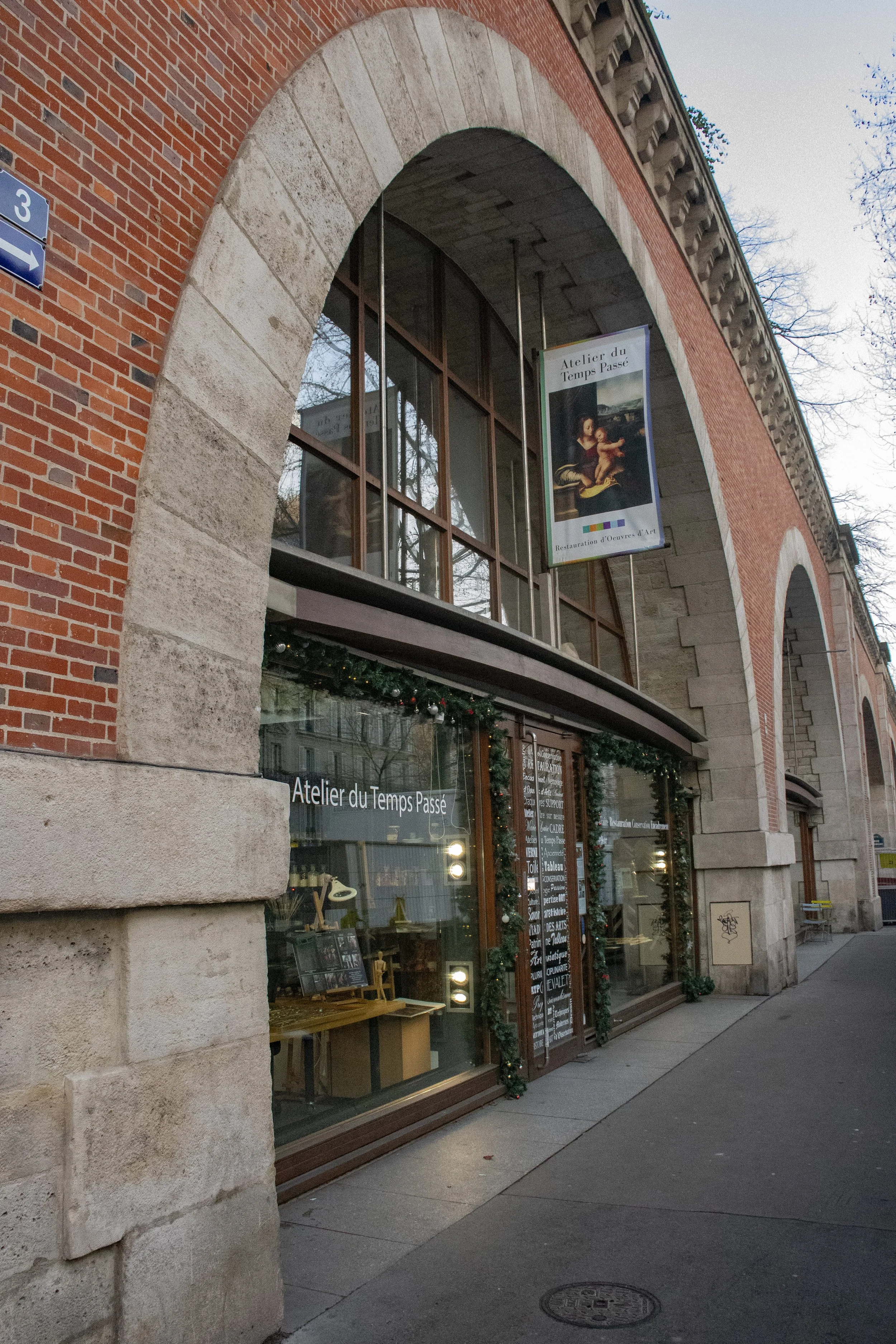 Façade d'un bâtiment en briques avec un grand arc en pierre, une vitrine décorée avec une guirlande de Noël et un panneau indiquant l'atelier du Temps Passé, un lieu dédié à la restauration d'œuvres d'art.