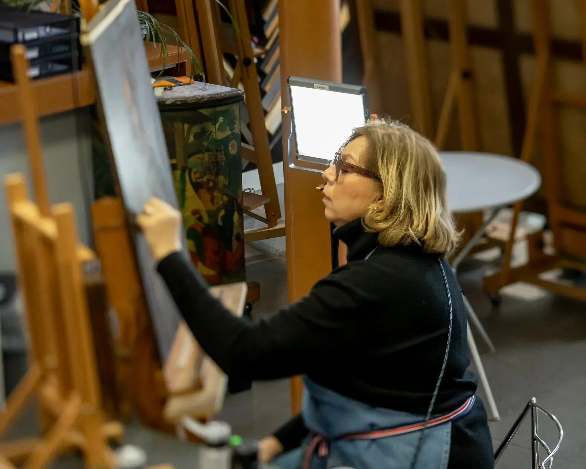Une femme peignant une œuvre d'art sur une toile dans un atelier, portant des lunettes et un tablier.