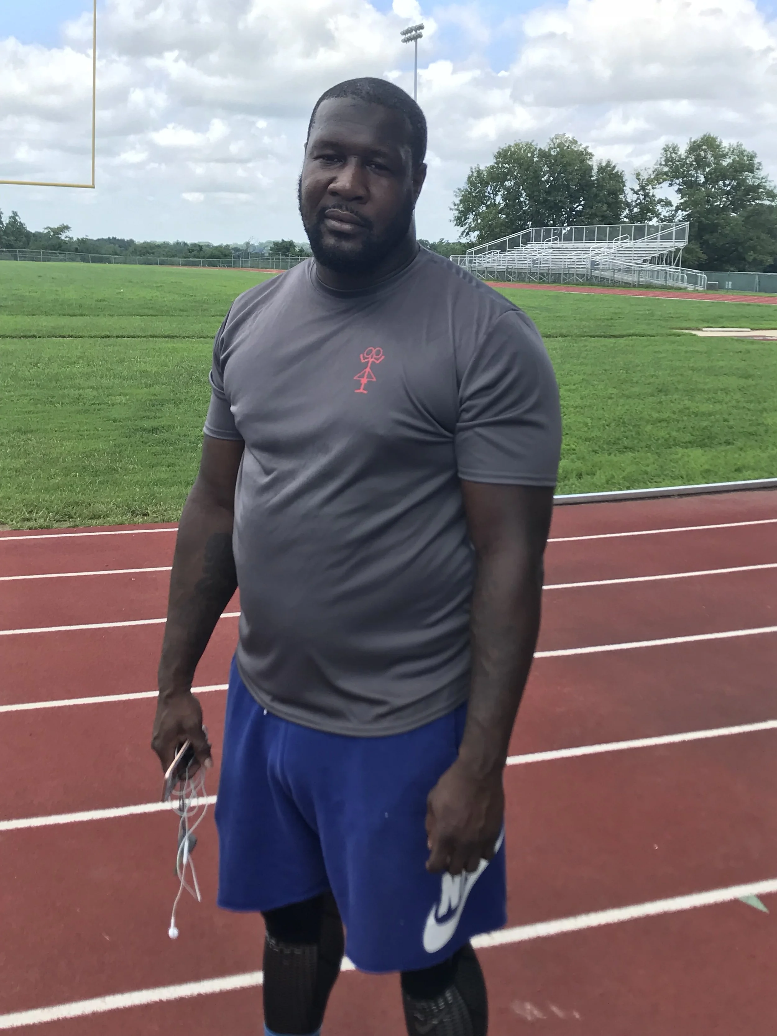 A man standing on a track field holding a phone and earphones, wearing athletic clothing, with green grass and trees in the background.