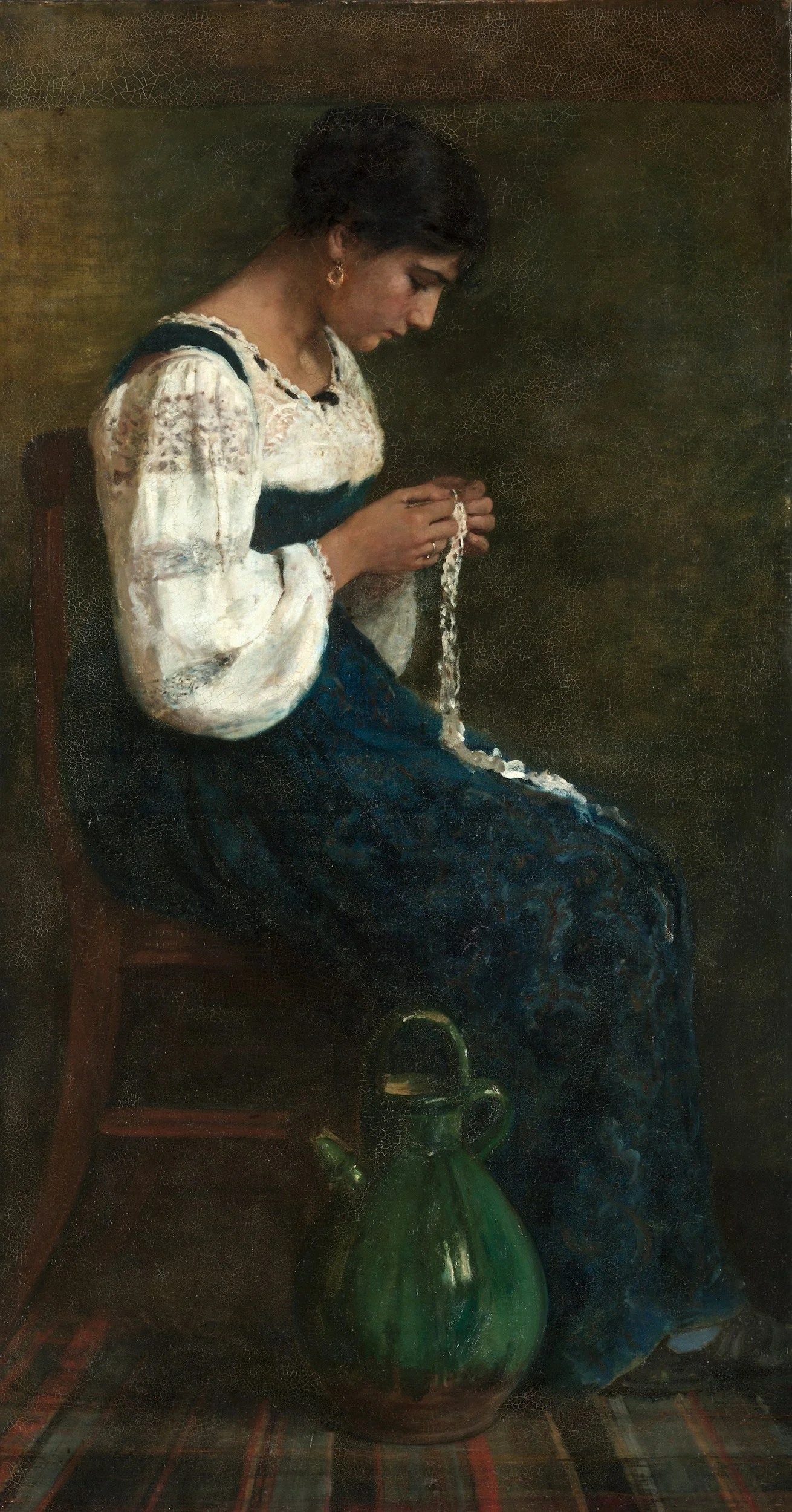 A woman sitting on a chair, looking down at a string of beads she is holding, with a green glass jug on the floor in front of her.