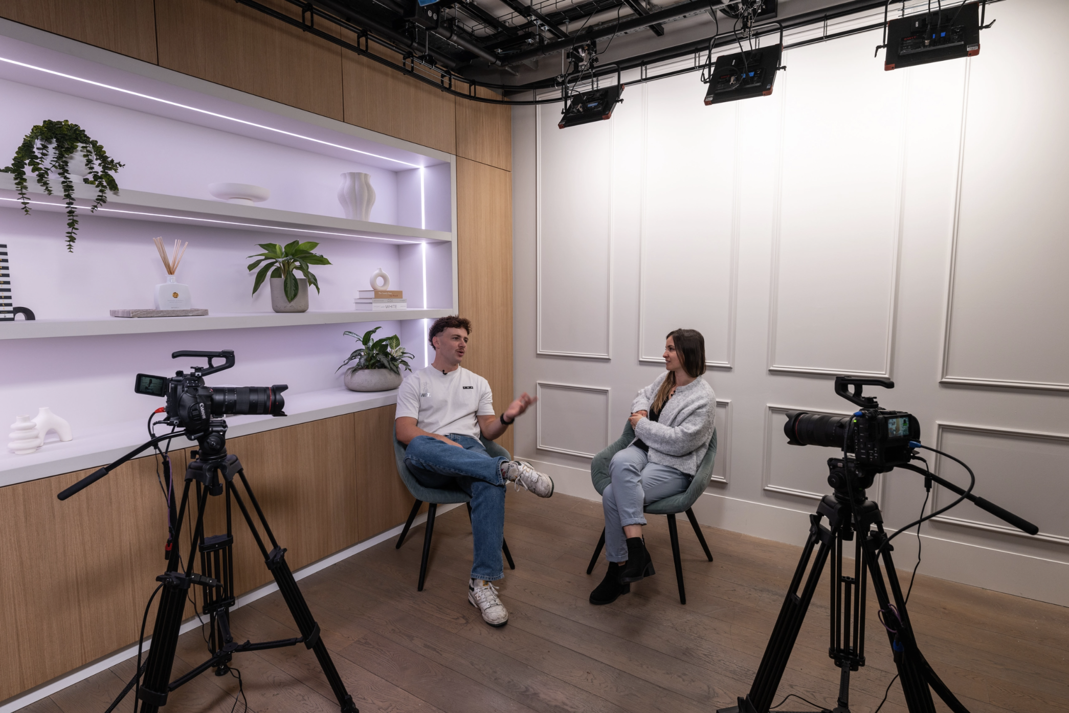 Two people speaking in a podcast studio with shelving and paneling behind them