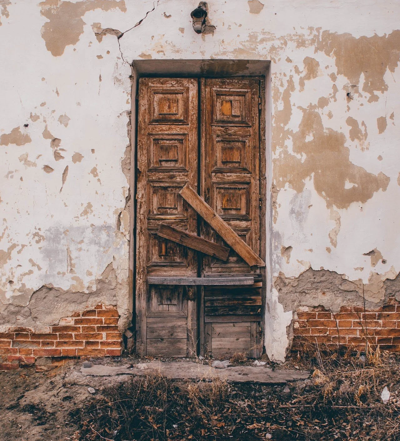 An old, wooden door with a broken and boarded-up section on a weathered, peeling wall with exposed bricks underneath.