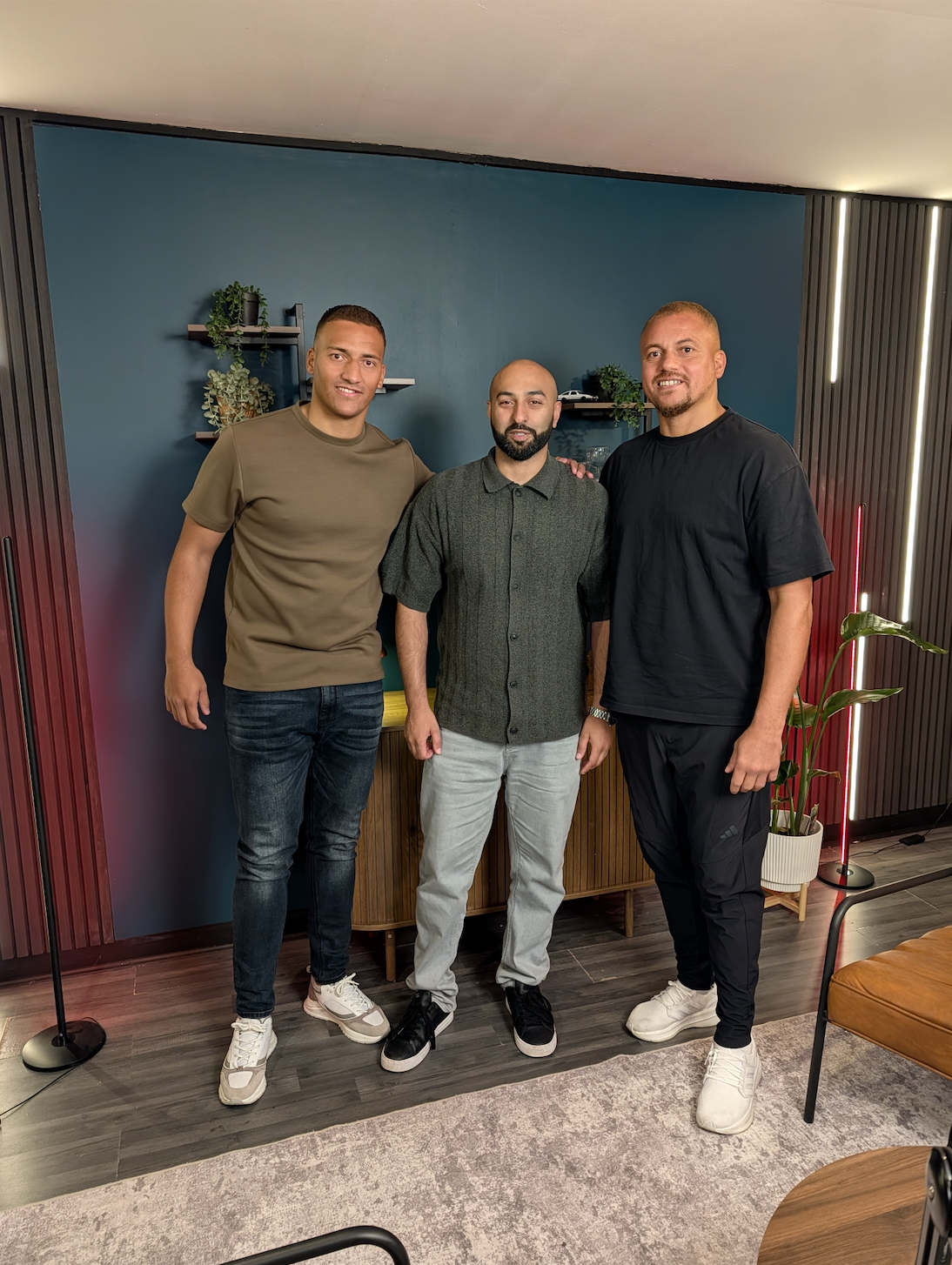 Three men standing together at Ancoats Podcast Studio in front of a teal wall with small shelves and plants, smiling at the camera.