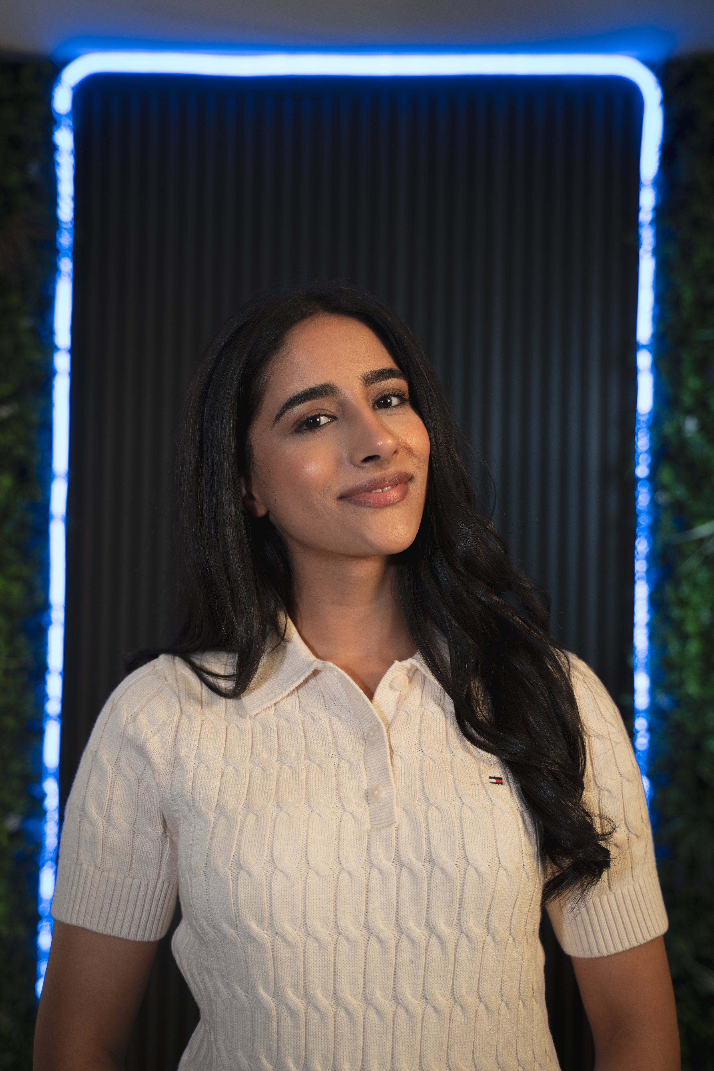 A young woman with long dark hair, wearing a cream-colored cable knit polo shirt, standing in front of a dark background with blue neon lighting at the edges. Taken at Ancoats Podcast Studio in Manchester.