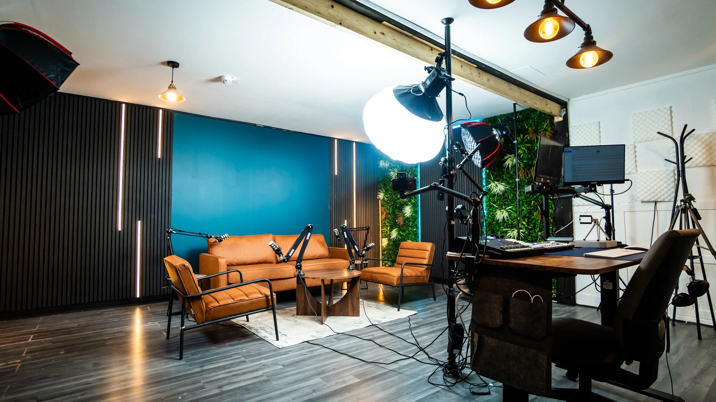 Ancoats Podcast Studio setup with a brown couch and chairs, professional lighting, cameras, monitors, and a green wall with plants.