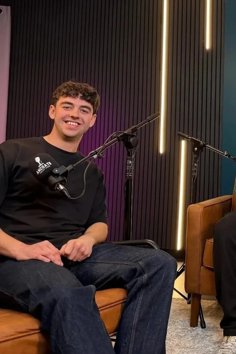 A young man with short curly hair smiling while sitting on a brown leather chair in Ancoats Podcast Studio, with microphones and black acoustic wall paneling in the background.