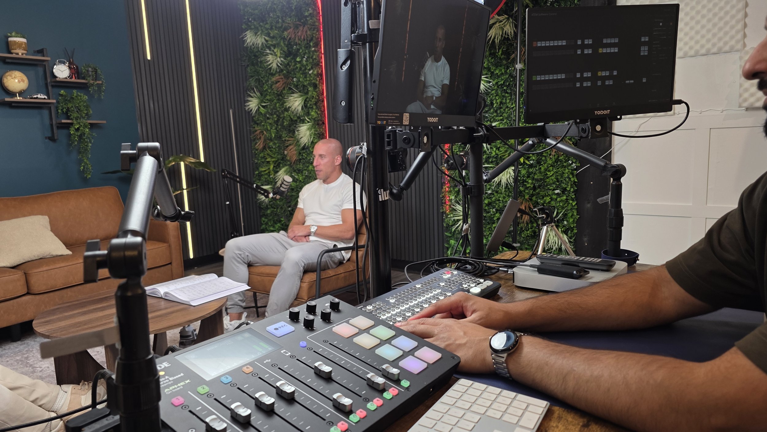 Video production studio with a man being recorded sitting on a brown sofa, microphones, monitors, and a control board in the foreground. Taken at Ancoats Podcast Studio in Manchester.