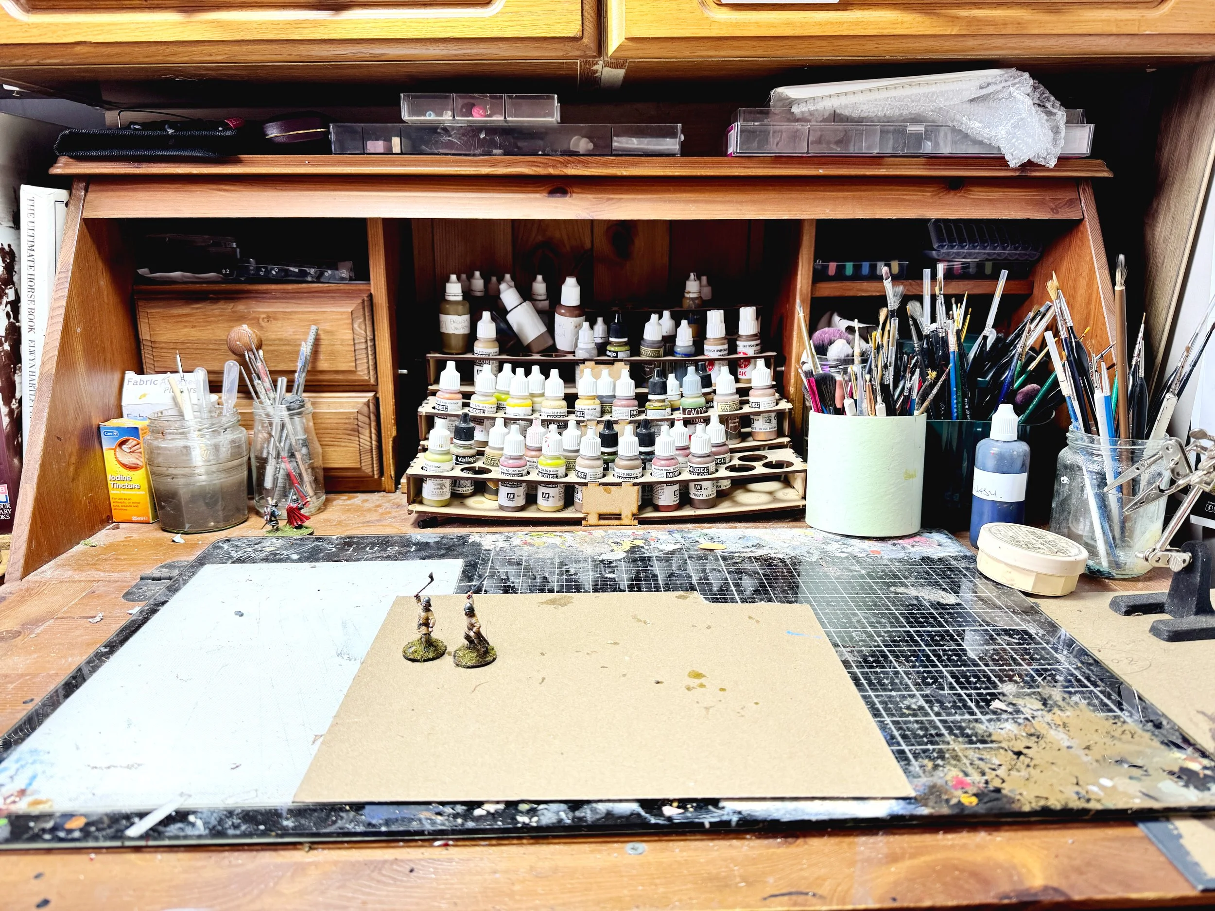 A tabletop artist's workspace with miniature figurines, paint bottles, brushes, and art supplies organized in jars and containers.