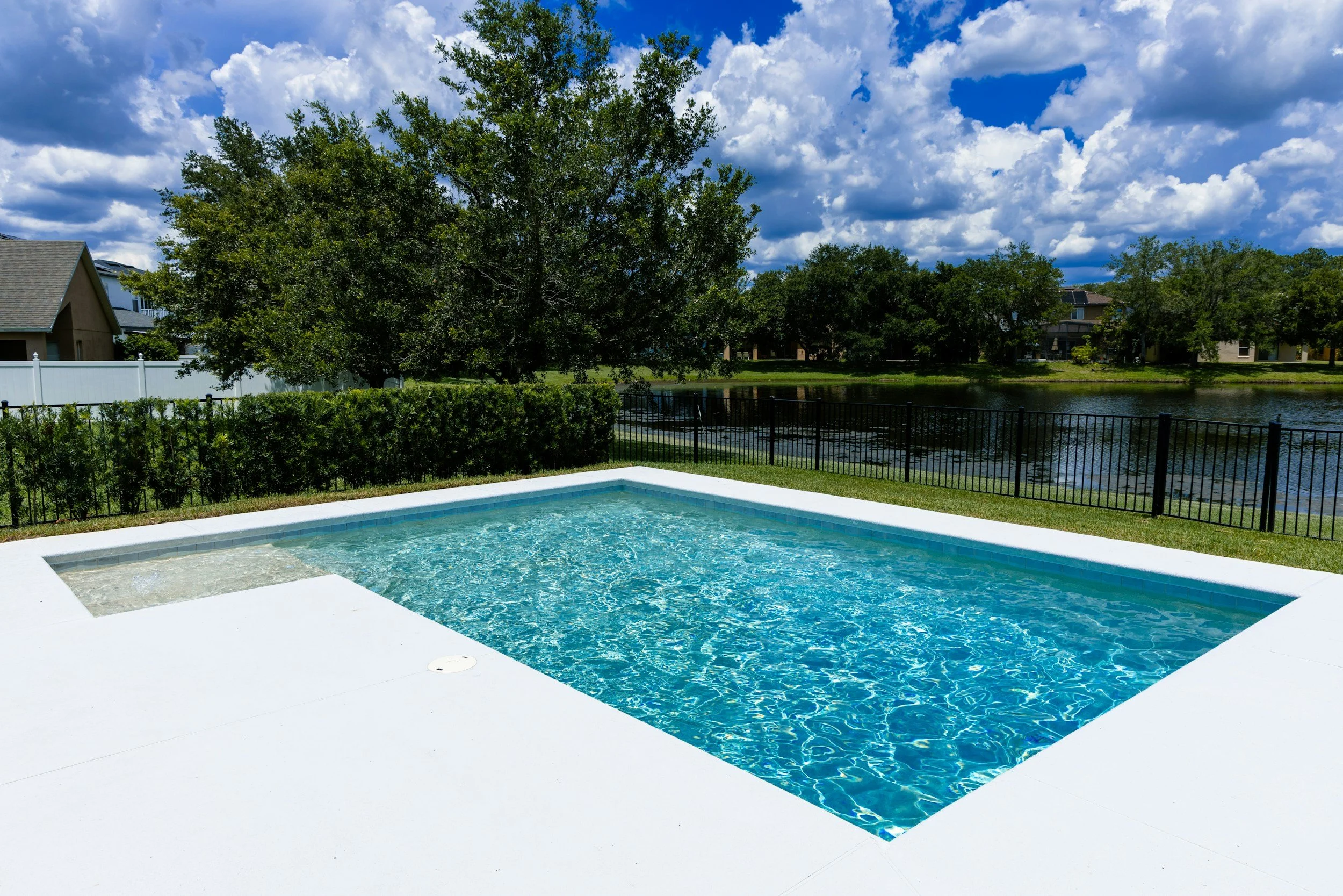A backyard pool with a white deck, surrounded by greenery and a black fence, overlooking a lake with houses and trees, under a partly cloudy sky.
