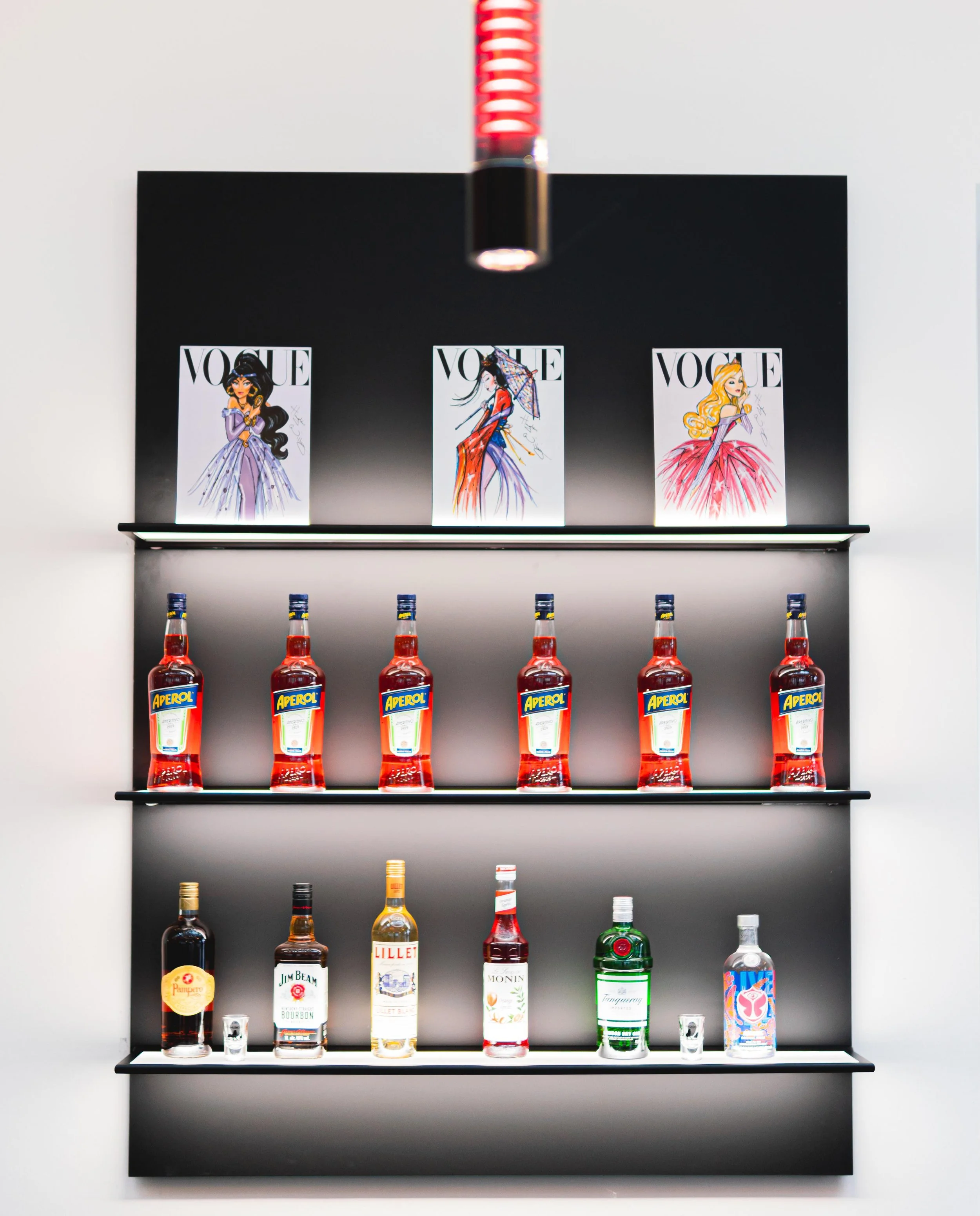 Regal mit Vodka und Aperol-Flaschen vor einer schwarzen Wand mit Vogue-Postern und einem roten Licht über dem Regal.