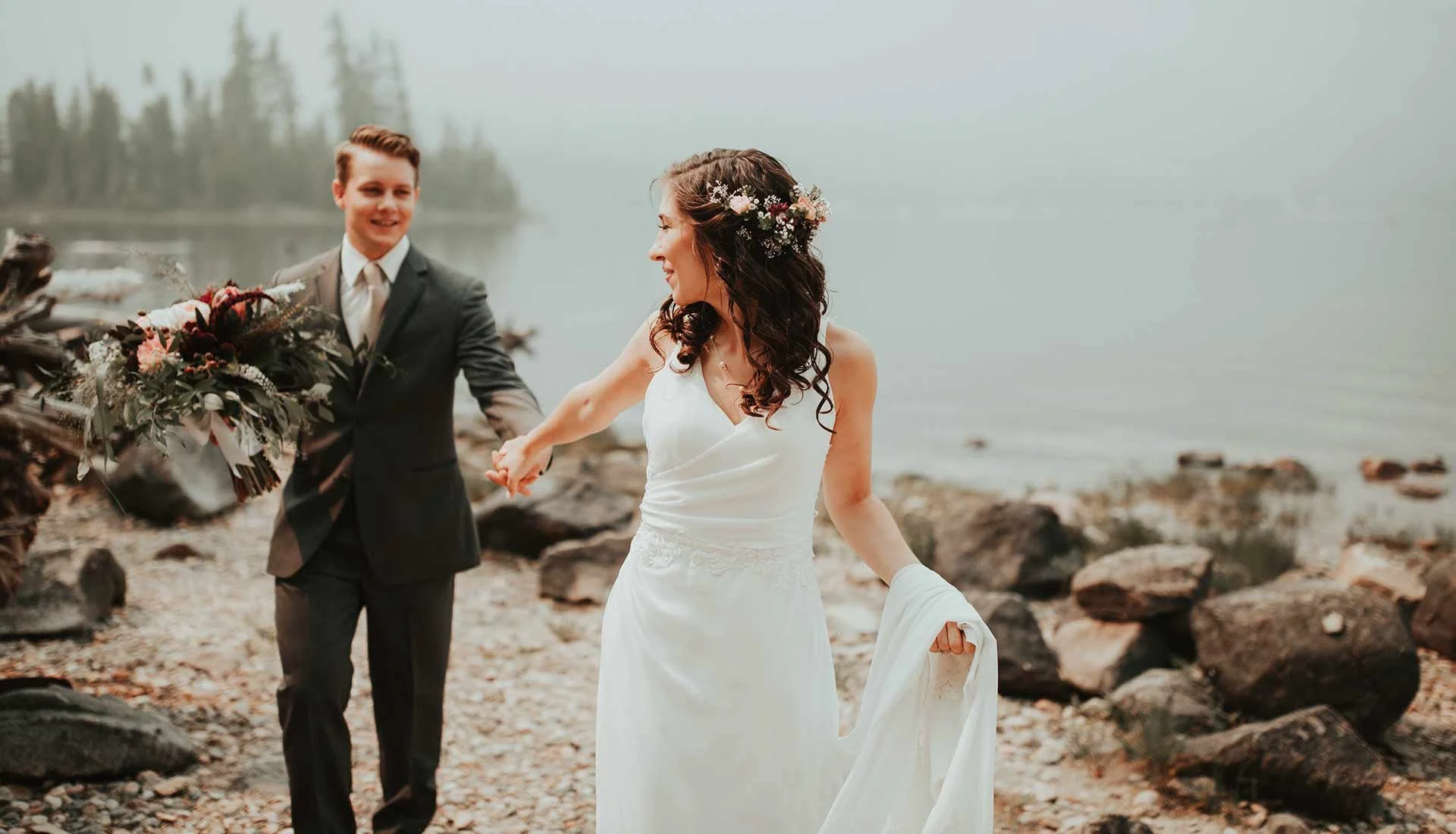 Ein Brautpaar, das am Ufer eines Sees tanzt, die Braut trägt ein weißes Kleid mit Blumen im Haar, der Bräutigam trägt einen schwarzen Anzug.