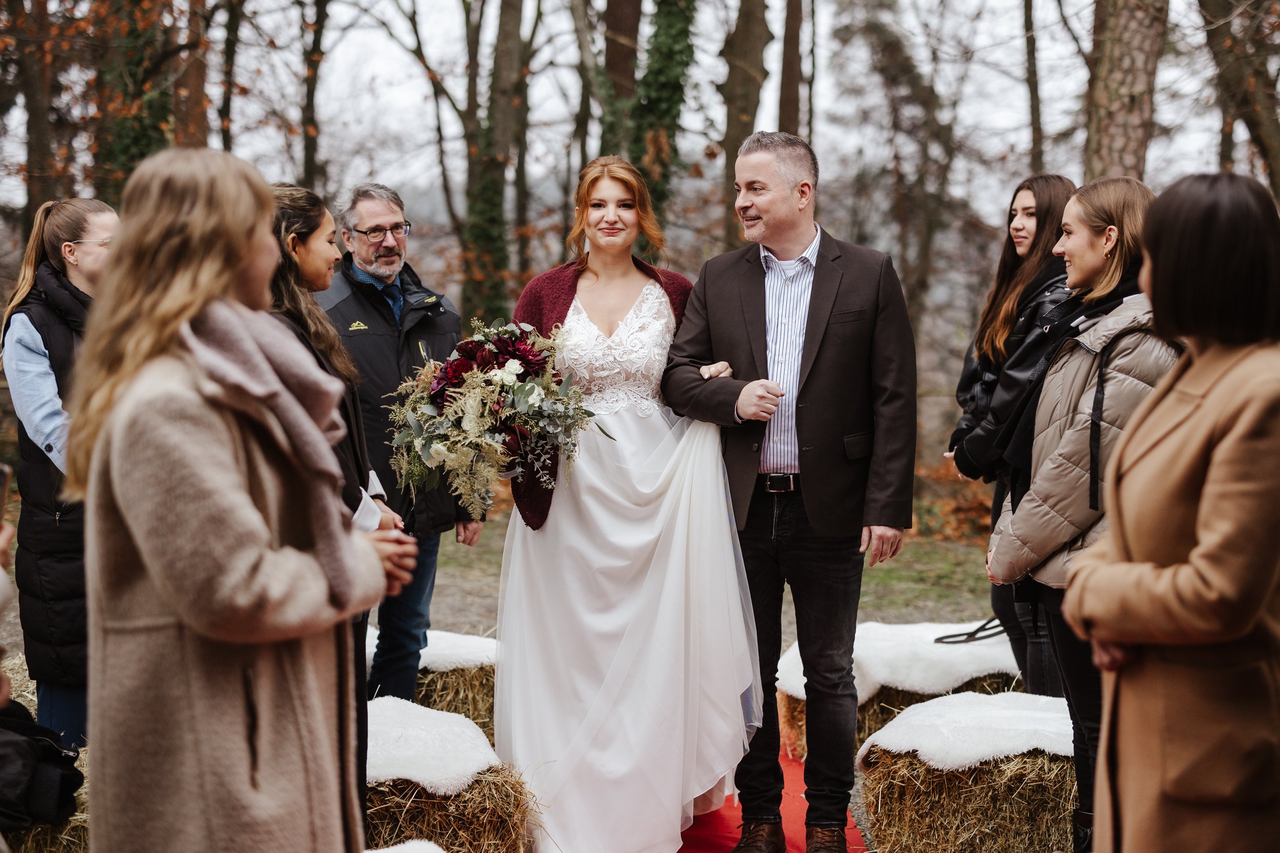 Vater führt die Braut zum Altar, umgeben von Freunden und Familie, auf einer Winterhochzeit im Wald.