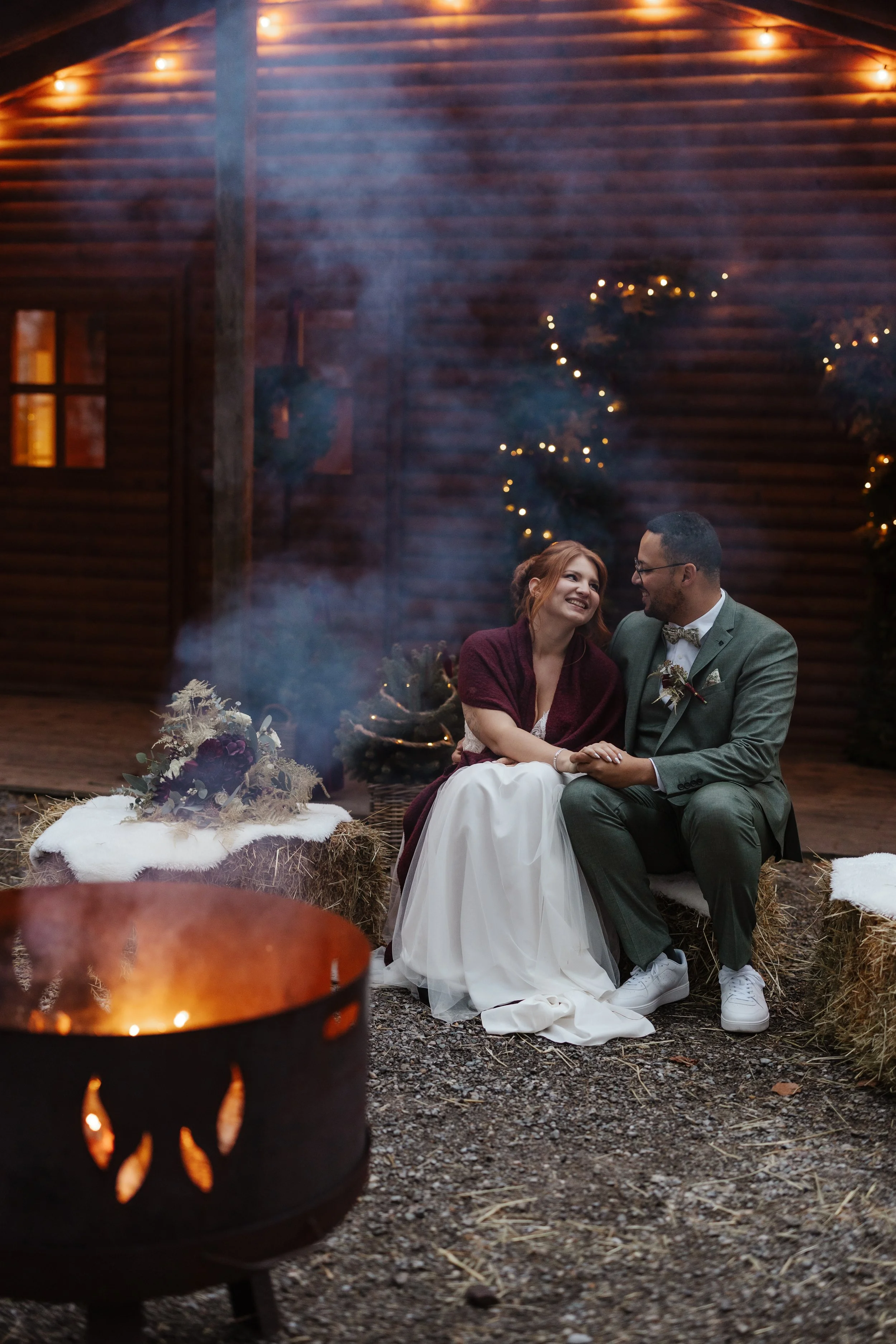Ein lachendes Brautpaar am Lagerfeuer, umgeben von Weihnachtsdeko und einem Feuerkorb bei winterlicher Atmosphäre.