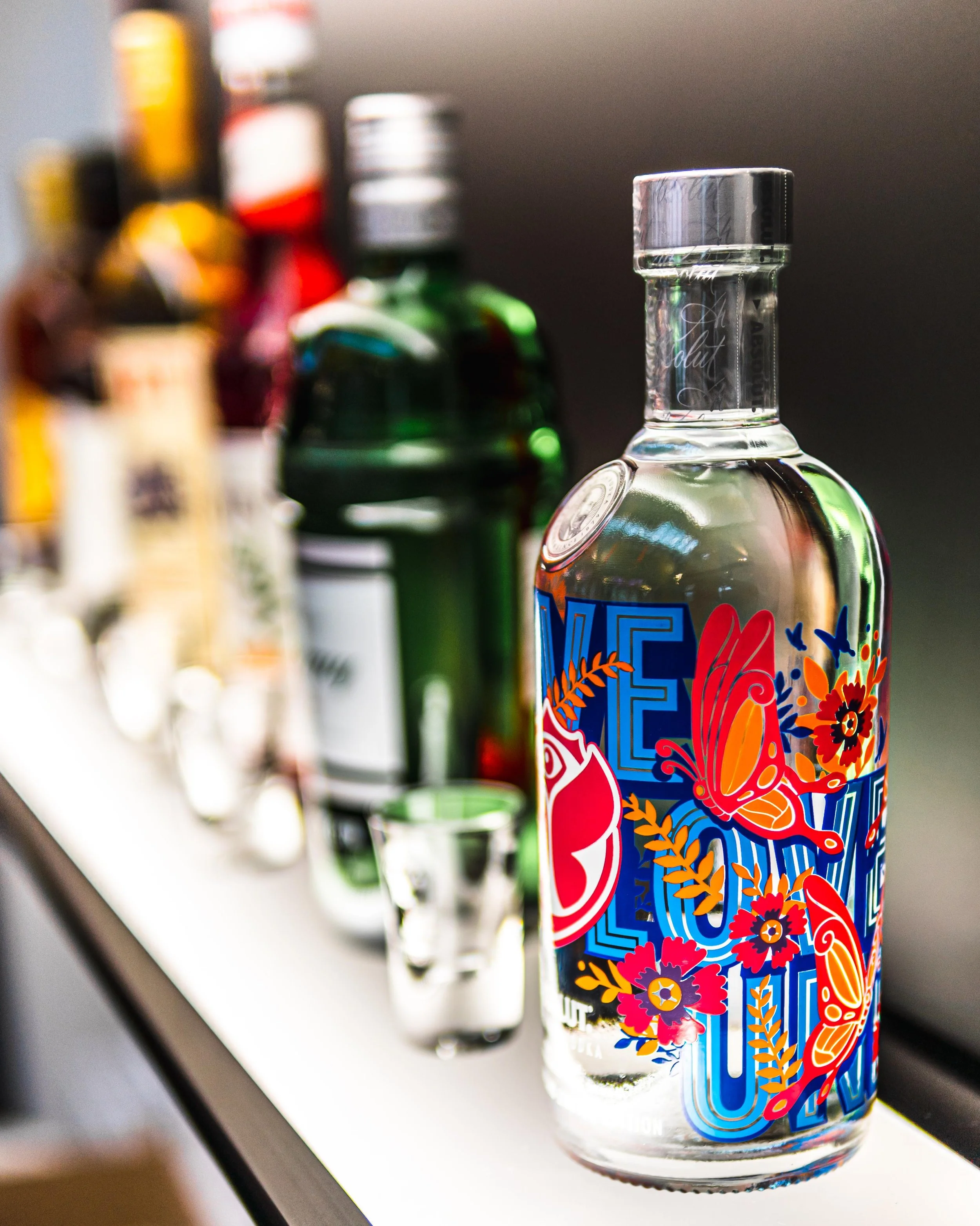 Eine Flasche mit buntem Blumen- und Schmetterlingsdesign auf der Vorderseite. Hinter der Flasche sind weitere Flaschen unterschiedlicher Farben und Formen, sowie ein kleiner Schnapsglas auf einer weißen Oberfläche. Der Hintergrund ist unscharf.