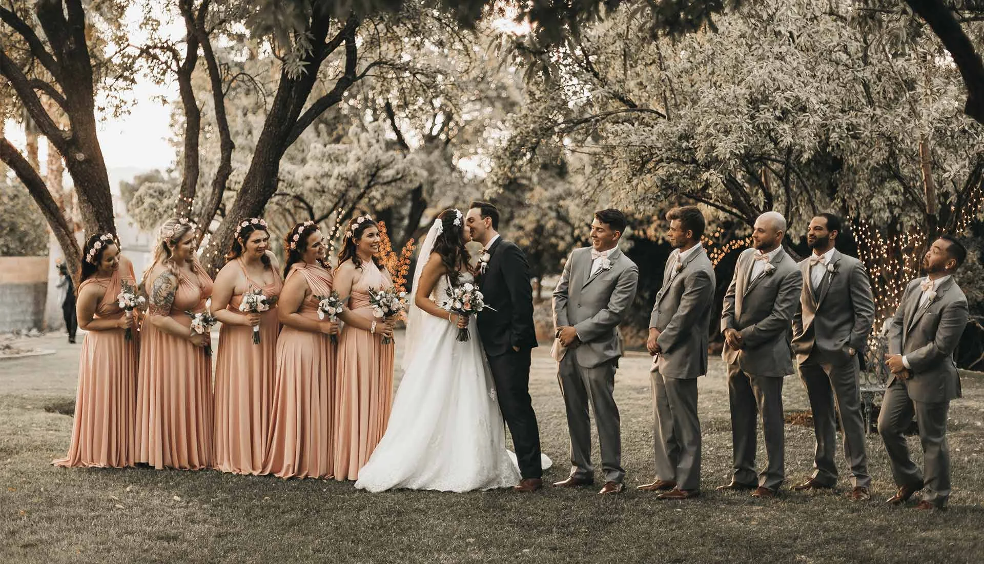 Ein Brautpaar küsst sich in der Mitte, umgeben von Brautjungfern in peachfarbenen Kleidern auf der linken Seite und Trauzeugen in grauen Anzügen auf der rechten Seite, im Freien unter Bäumen mit Lichterketten bei Sonnenuntergang.
