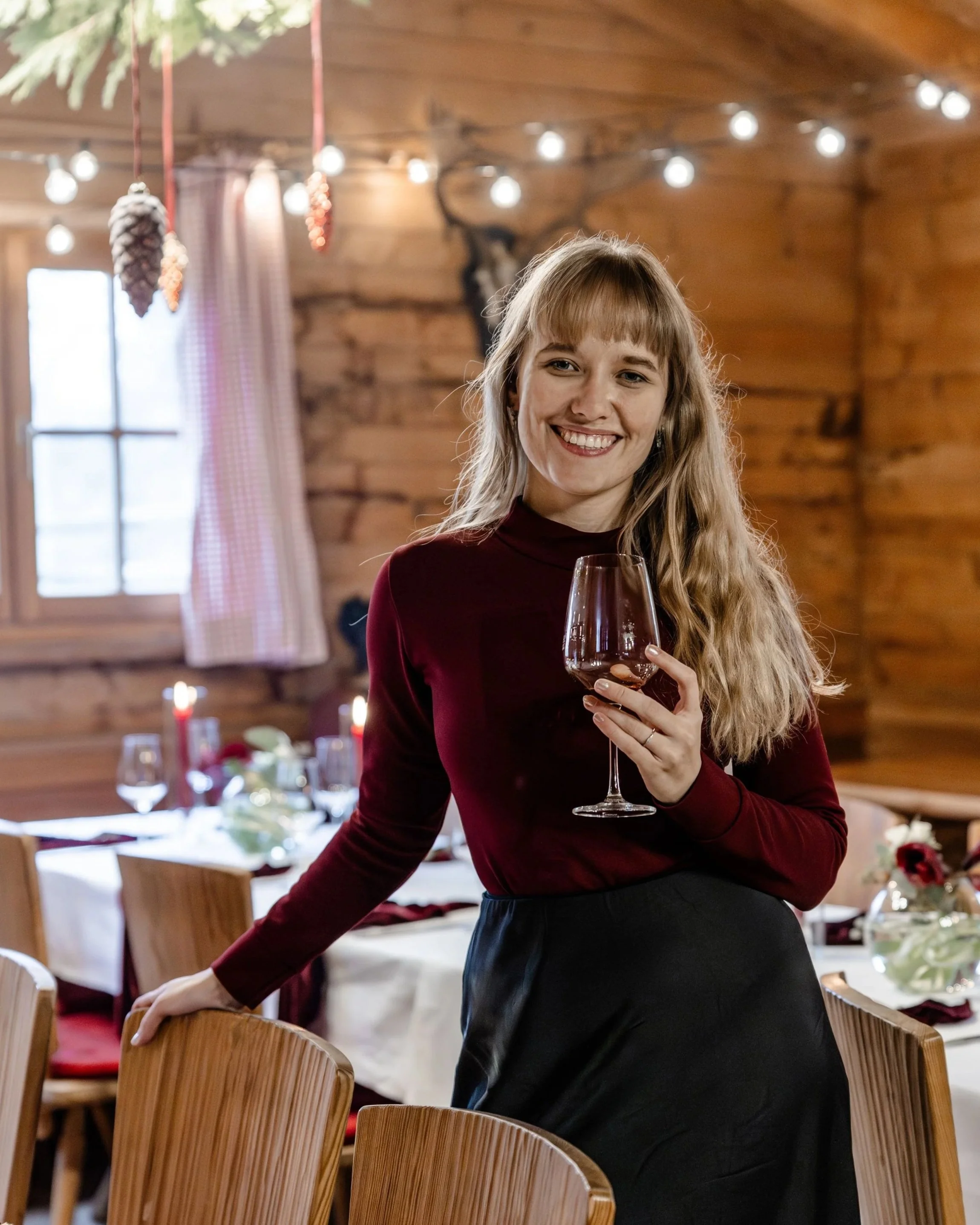 Hochzeitsplanerin bei einer Hochzeitsfeier, die ein Weinglas hält, steht in einem rustikalen Holzintérieur mit festlicher Dekoration und gedecktem Tisch im Hintergrund.