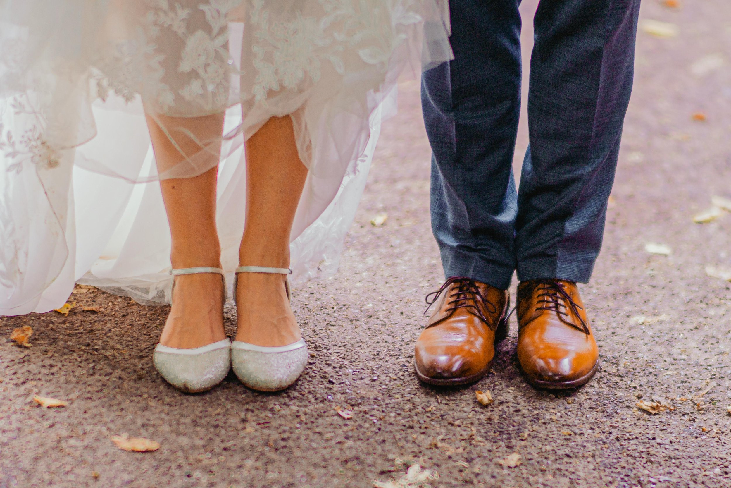 Nahaufnahme der Füße eines Paares bei einer Hochzeit, die weibliche Person trägt hochhackige Schuhe, der männliche Partner trägt braune Lederschuhe. Sie stehen auf einer Erde- und Laubbeschicht.