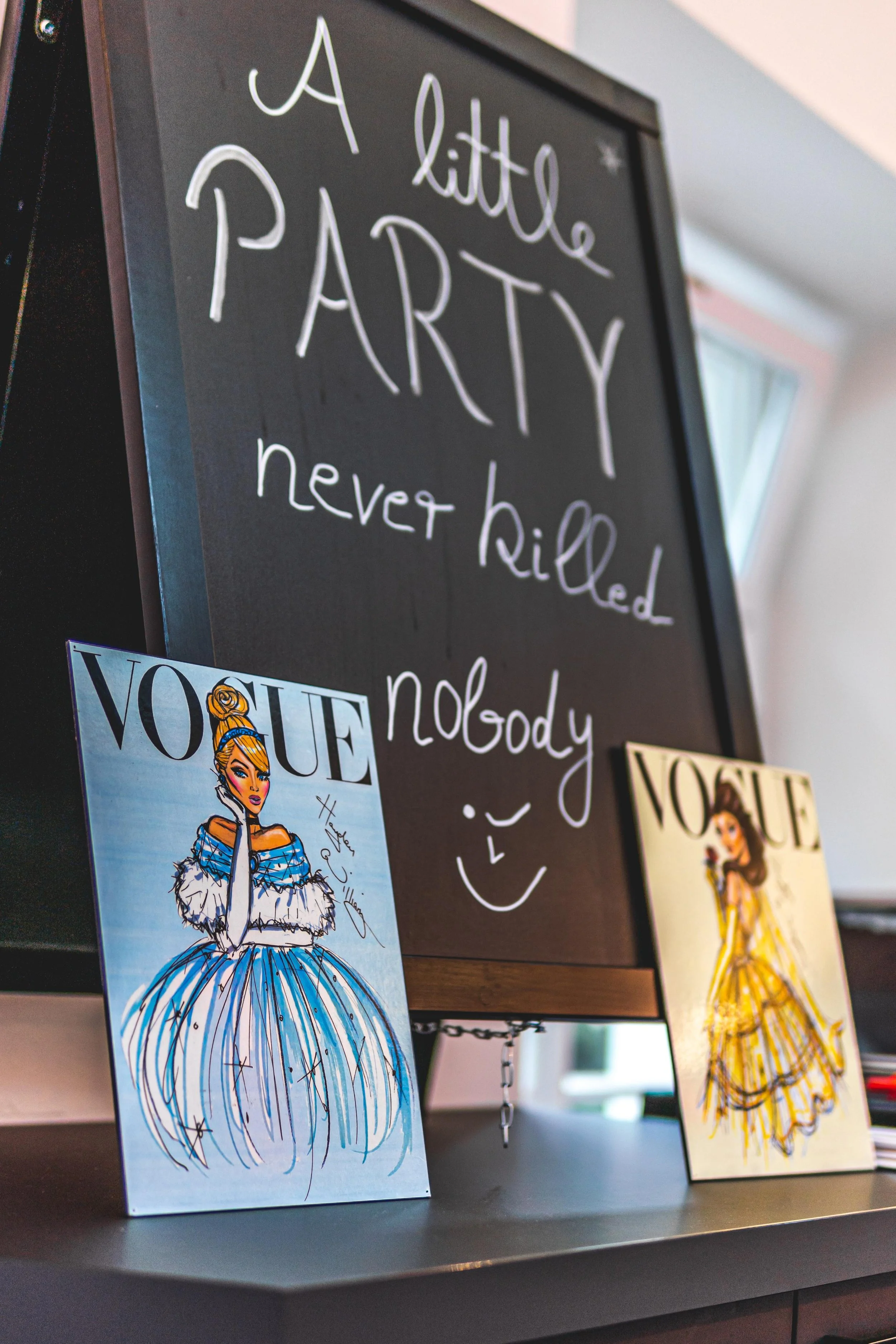 Eine schwarze Tafel mit weißen handgeschriebenen Worten: "A little PARTY never killed nobody". Zwei Vogue-Magazine sind vor die Tafel gestellt, eine mit einer Zeichnung einer eleganten Frau in einem bunten Outfit und die andere mit einer Abbildung ei