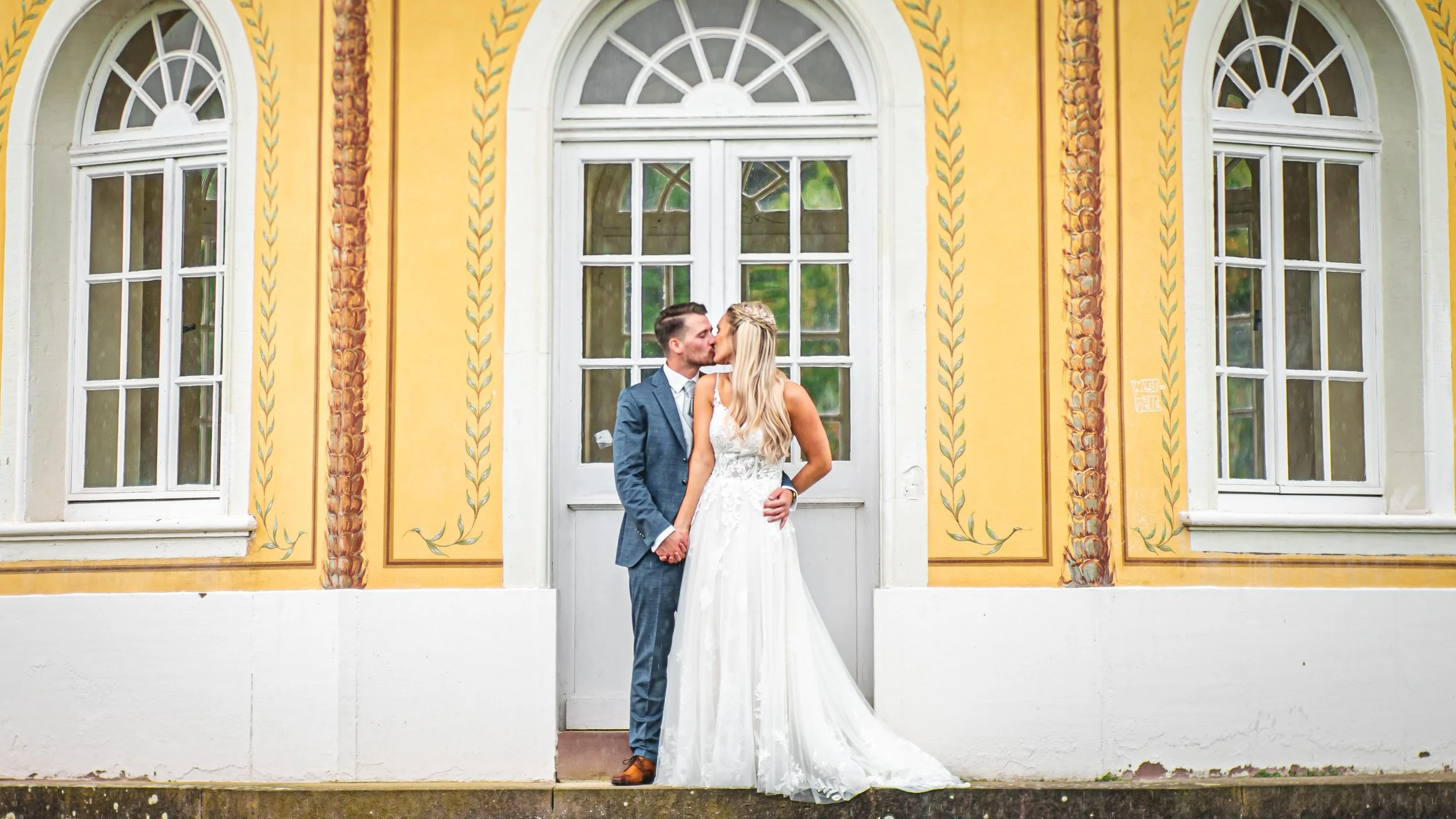 Braut und Bräutigam küssen sich vor einer bunten, dekorativen Hauswand mit großen Fenstern.