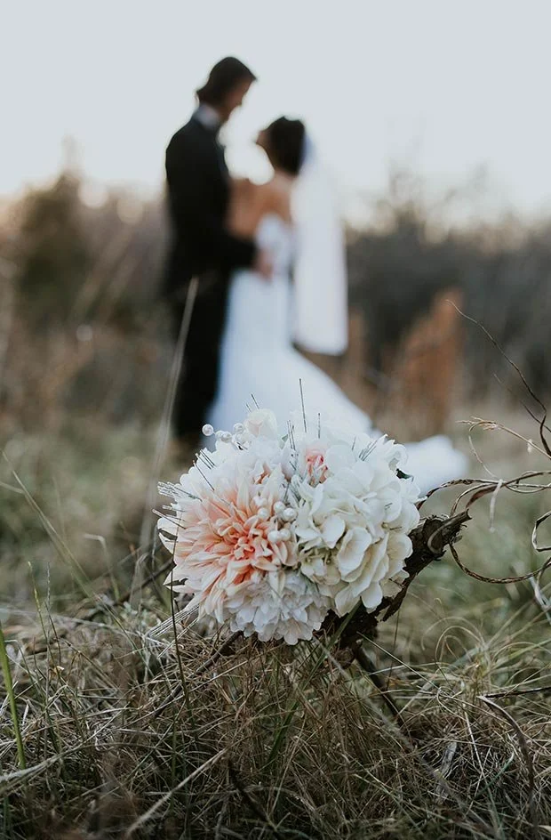 Verwackeltes Hochzeitsfoto mit Braut und Bräutigam im Hintergrund und Brautstrauß im Vordergrund in einer natürlichen Landschaft