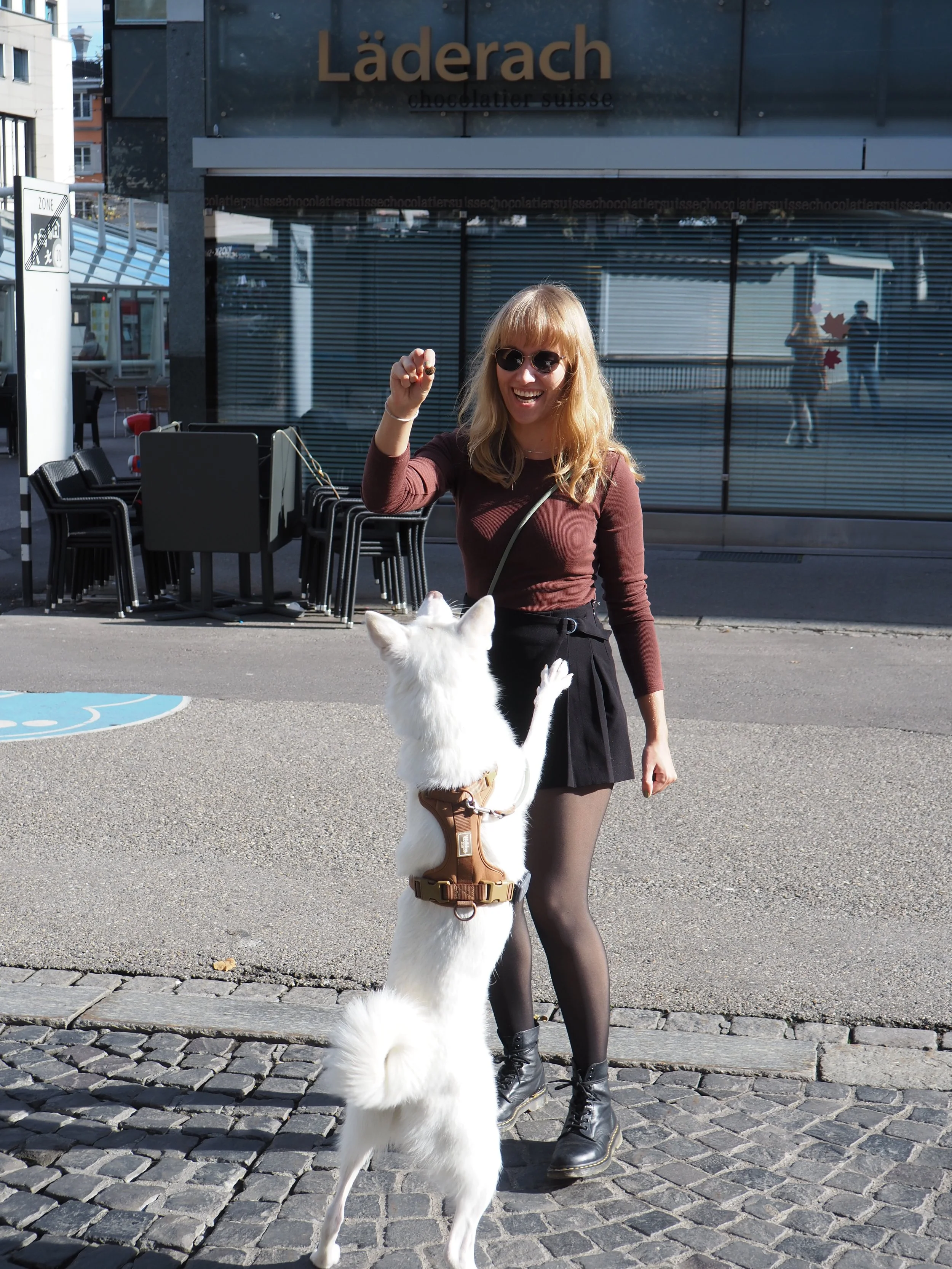 Hochzeitsplanerin Vanessa Schock mit Sonnenbrille beim Stadtspaziergang mit ihrer Hündin Luna, spielerisch vor einem Läderach-Geschäft – moderner Alltag zwischen Planung und Lebensfreude.