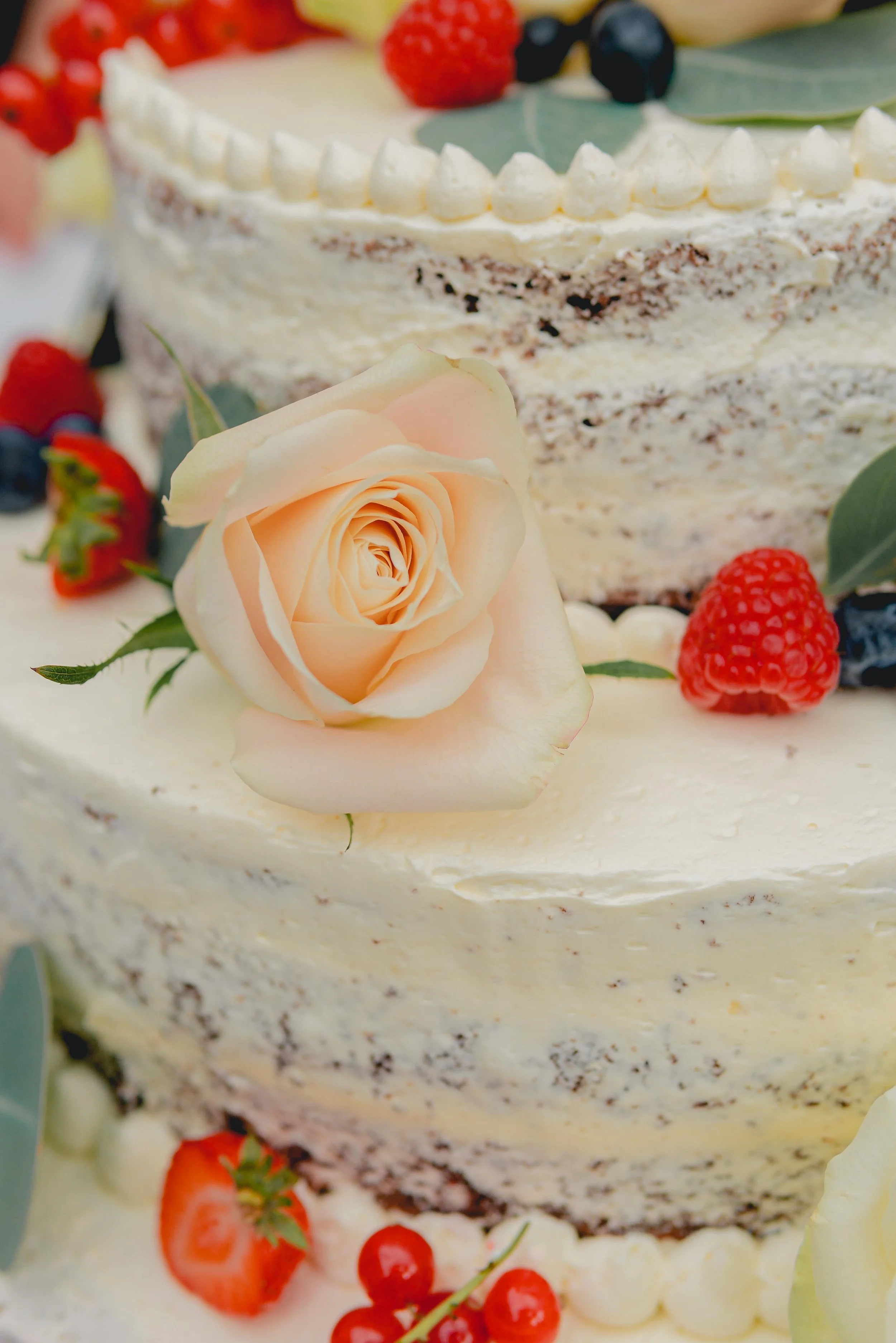 Nahaufnahme einer mehrstöckigen Torte mit weißer Creme, verziert mit einer weißen Rose, Erdbeeren, Himbeeren, Heidelbeeren und grünen Blättern.