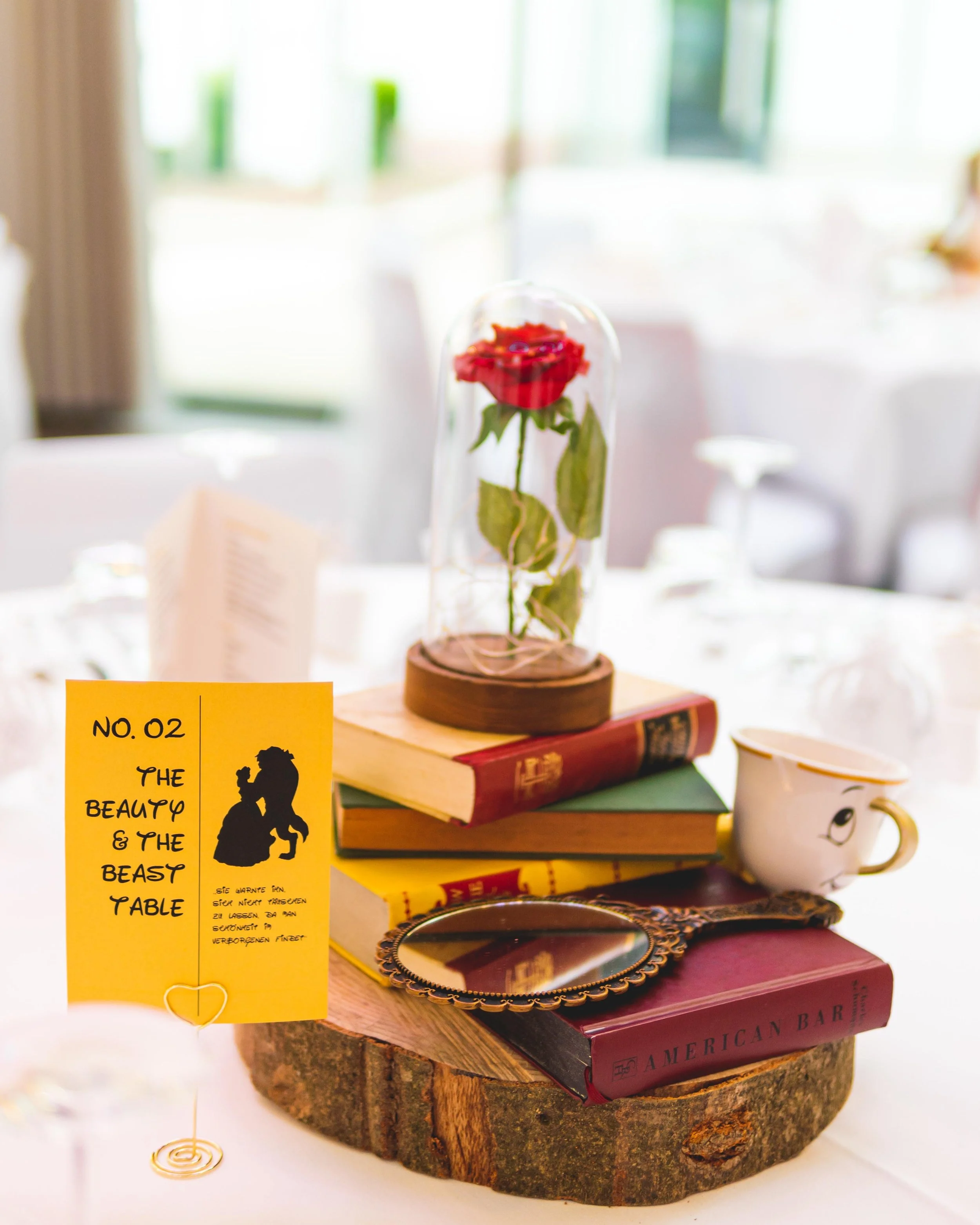 Tischdekoration mit verzauberten Büchern, einer Rose unter einer Glasscheibe, einem Kaffeebecher mit Gesicht, einem kleinen Spiegel, und einer gelben Karte mit einem Märchenbild und Text, auf einem Baumstumpf