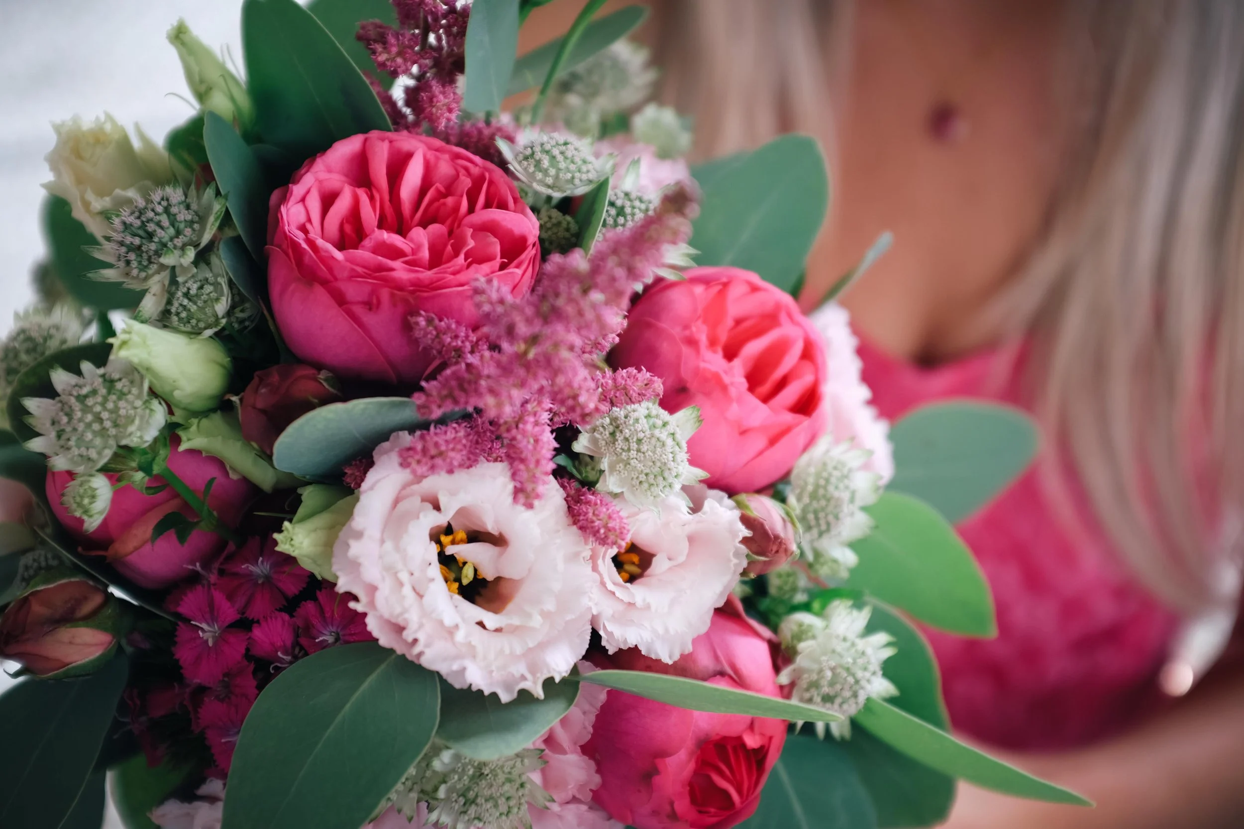 Nahaufnahme eines bunten Blumenstraußes mit rosa, weißen und lavendelfarbenen Blumen, umgeben von grünen Blättern, gehalten von einer Person in rosa Kleidung.