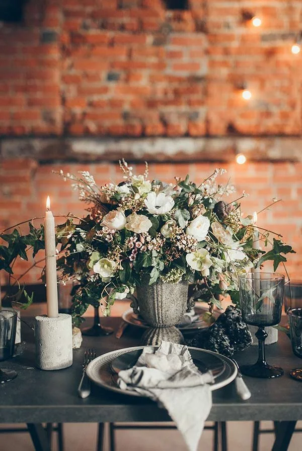 Stilvoll gedeckter Hochzeitstisch mit Blumen, Kerze, Geschirr und Backsteinwand – elegantes Hochzeitsdetail für Feiern im Bodenseeraum.