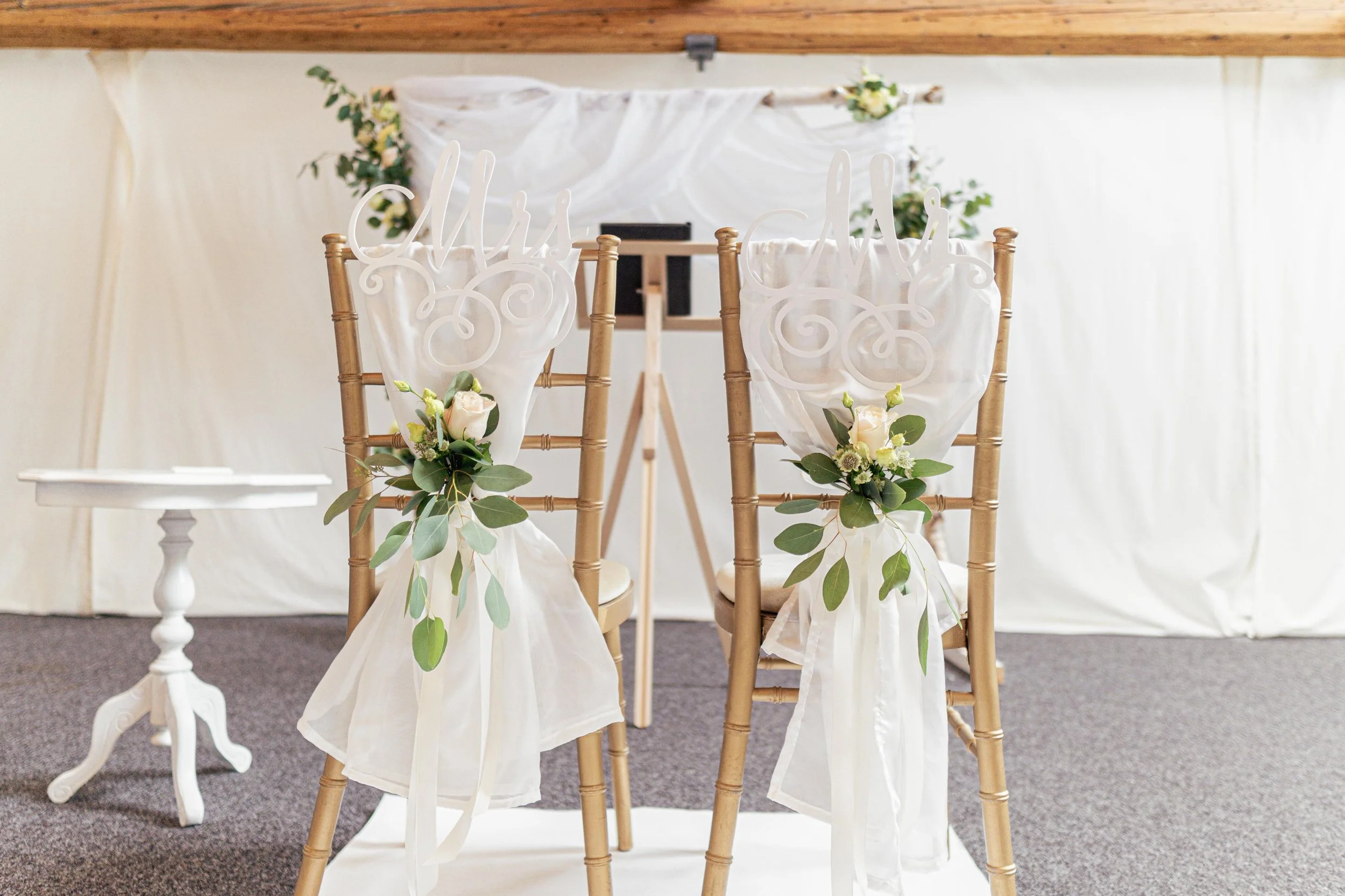 Zwei goldene Stühle mit weißen Tüchern, Blumen und "Mrs" und "Mr" Schild bei einer Hochzeitsfeier.