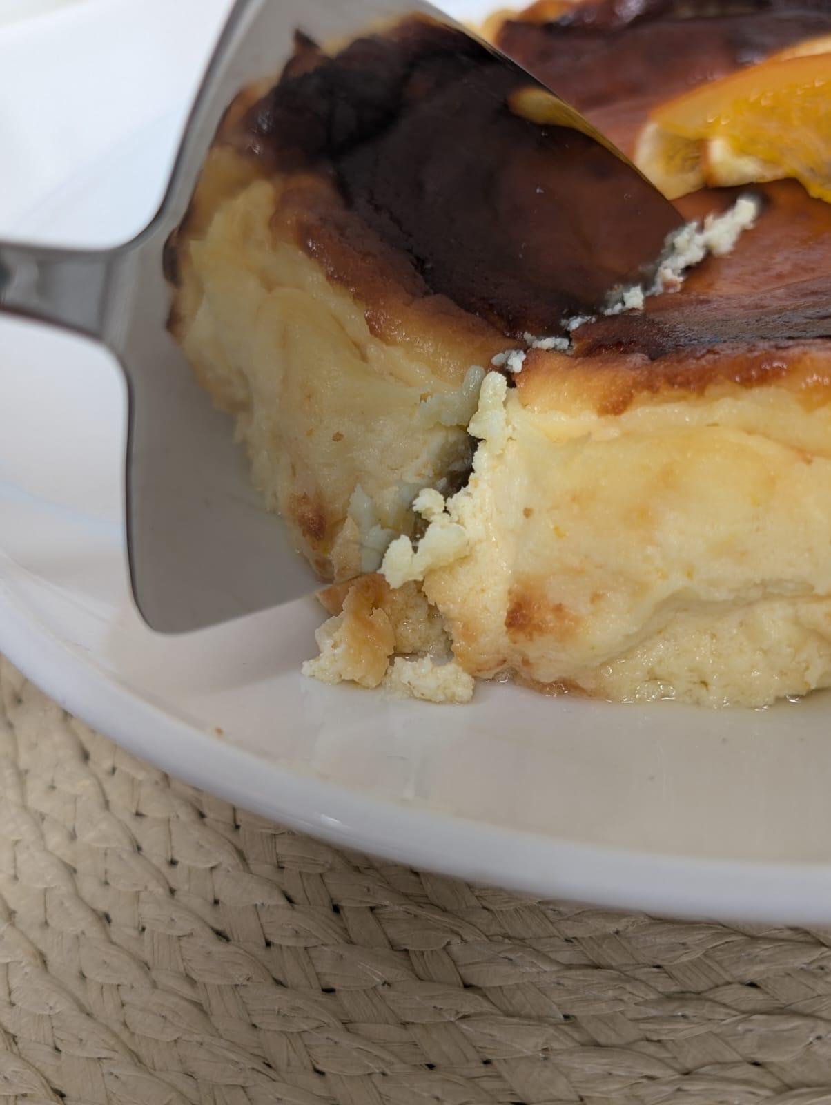 Close-up of a cheesecake being sliced with a metal spatula, topped with an orange slice, on a white plate.