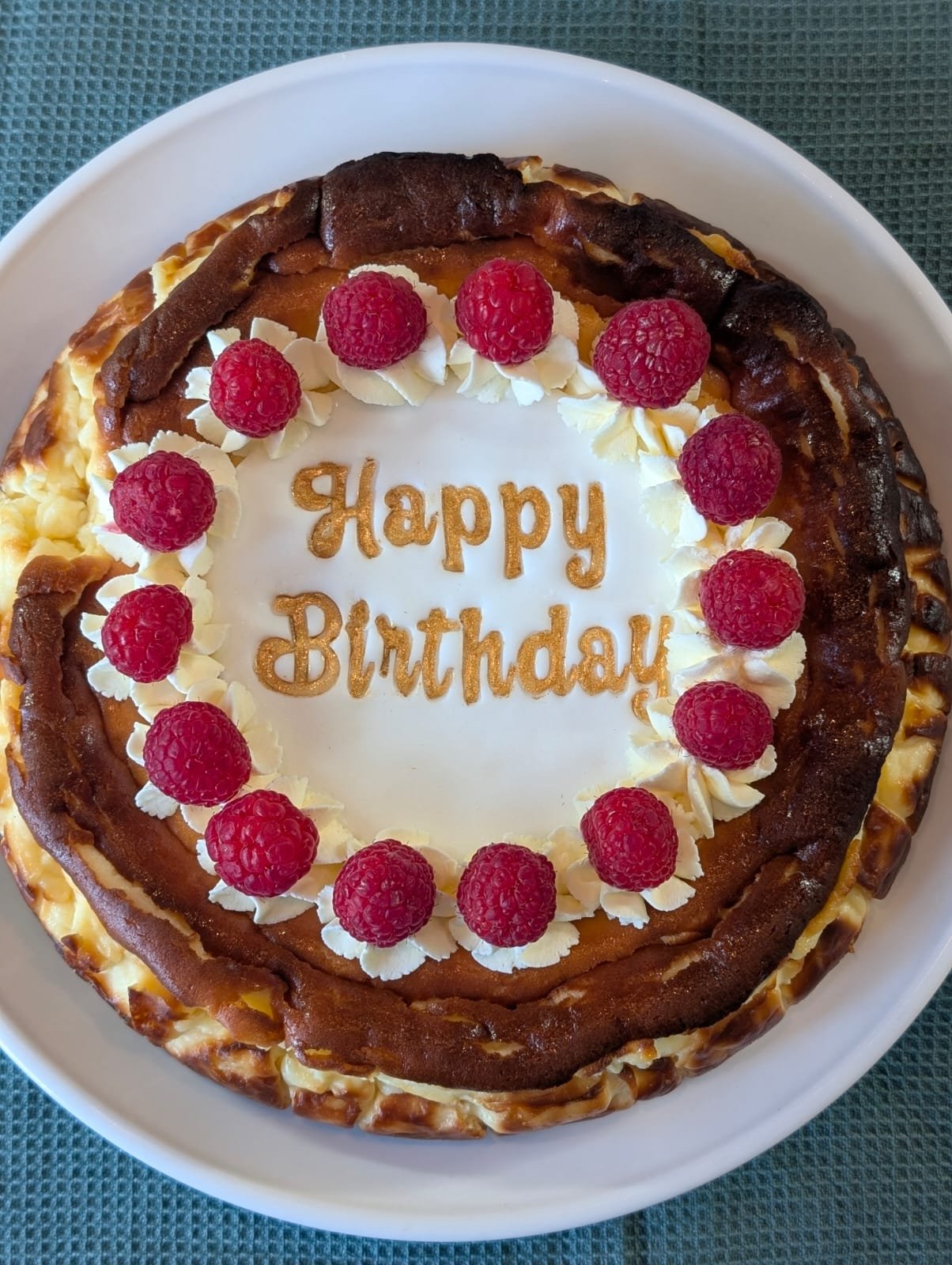 Cheese cake with "Happy Birthday" inscribed, surrounded by raspberries and cream, on a plate.
