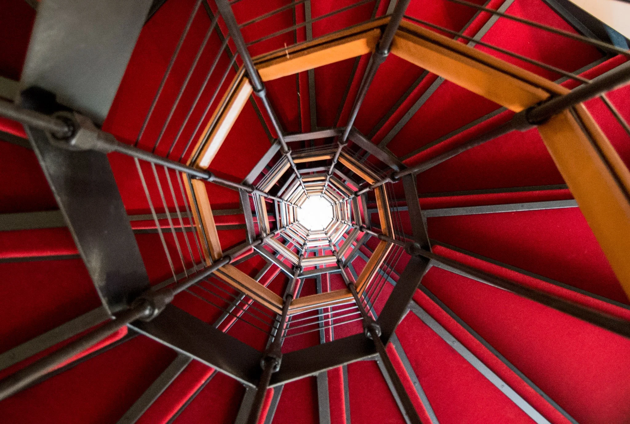 Looking up at a spiral staircase with red carpeted steps and black metal railings, converging towards a bright light at the top.