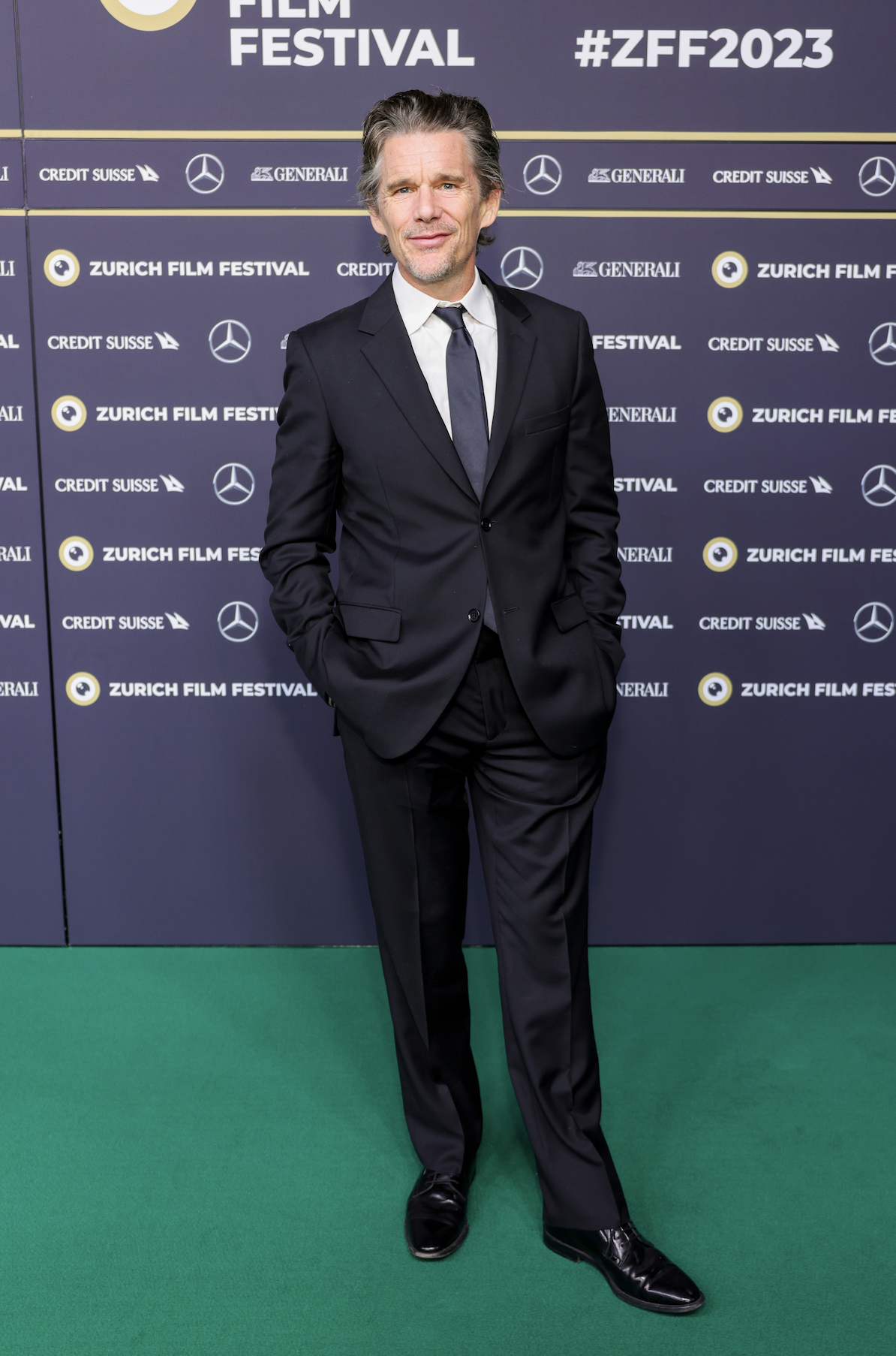 Man in a black suit standing on green carpet at Zurich Film Festival with a backdrop featuring event branding and sponsor logos.