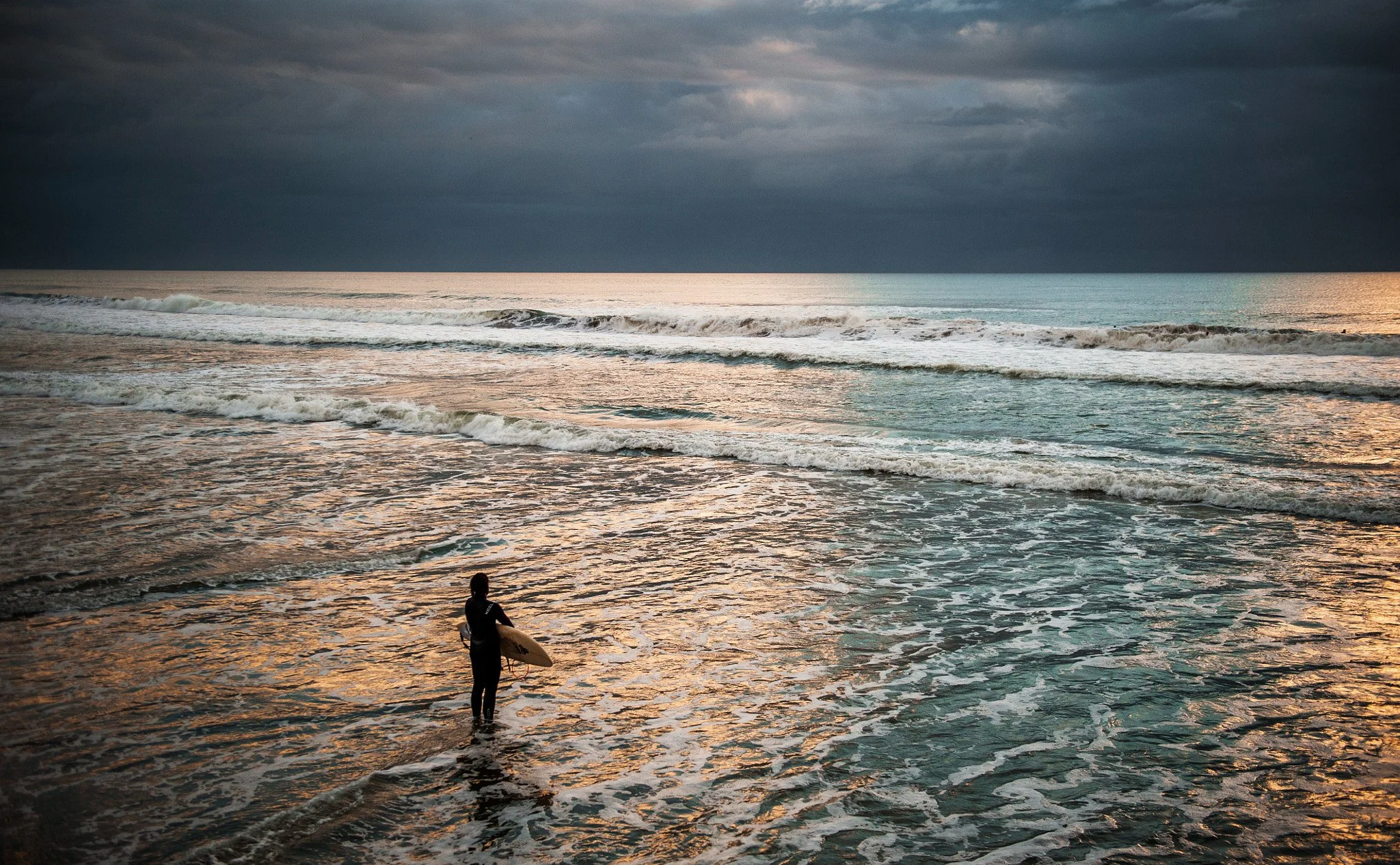 A person holding a surfboard standing at the edge of the ocean at sunset, with dark stormy clouds in the sky and gentle waves lapping at the shore.