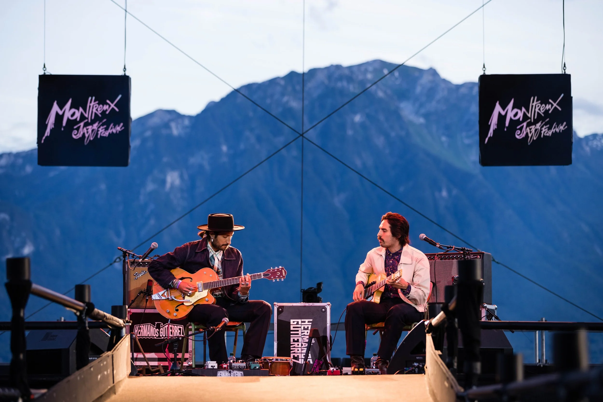 Two musicians perform guitar on a stage with a mountain backdrop, during Montreux Jazz Festival, with two lit signs reading 'Montreux Jazz Festival' hanging above.