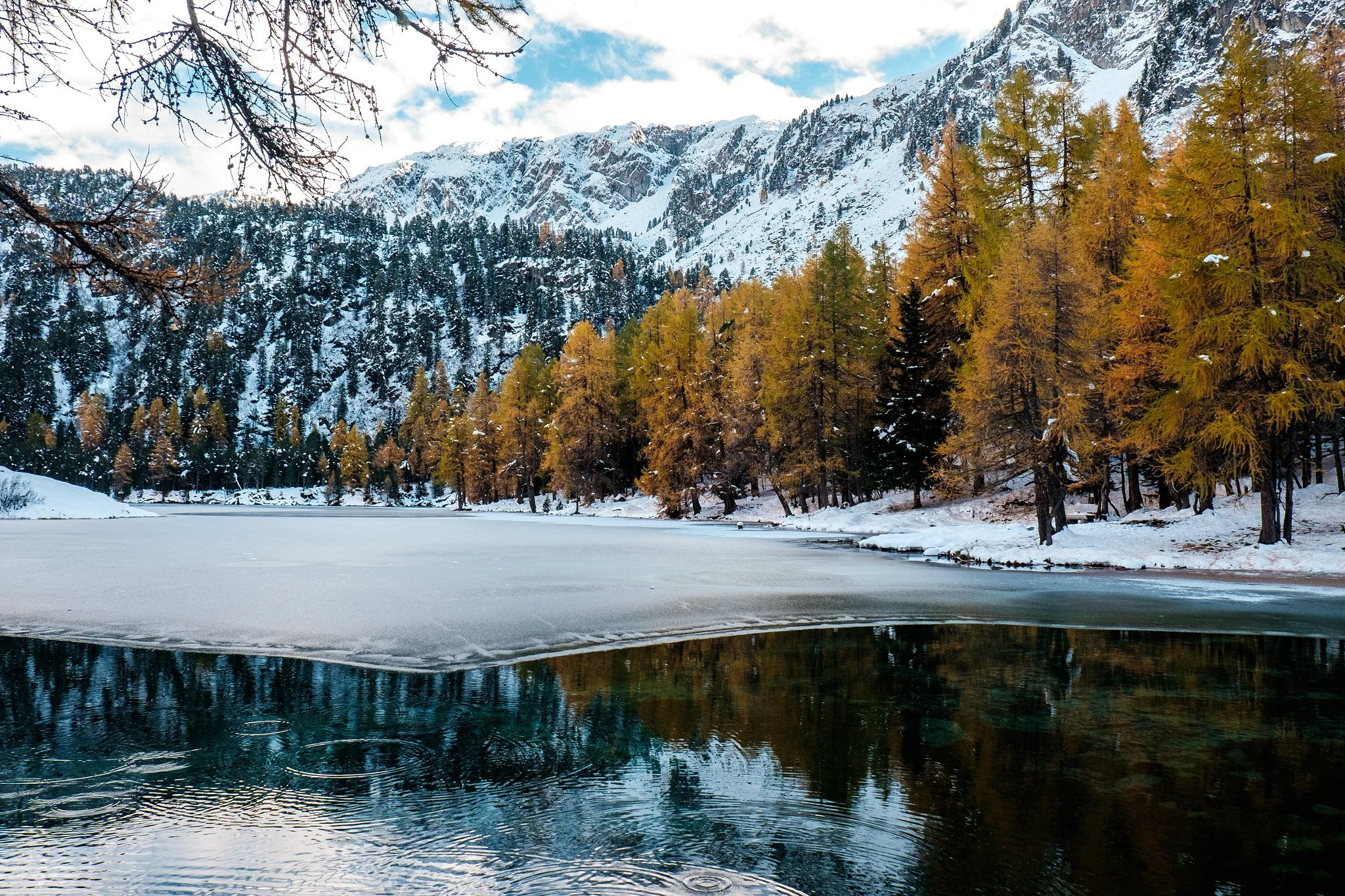 Snow-covered mountains and a partially frozen lake with trees along the shoreline, in a winter landscape.