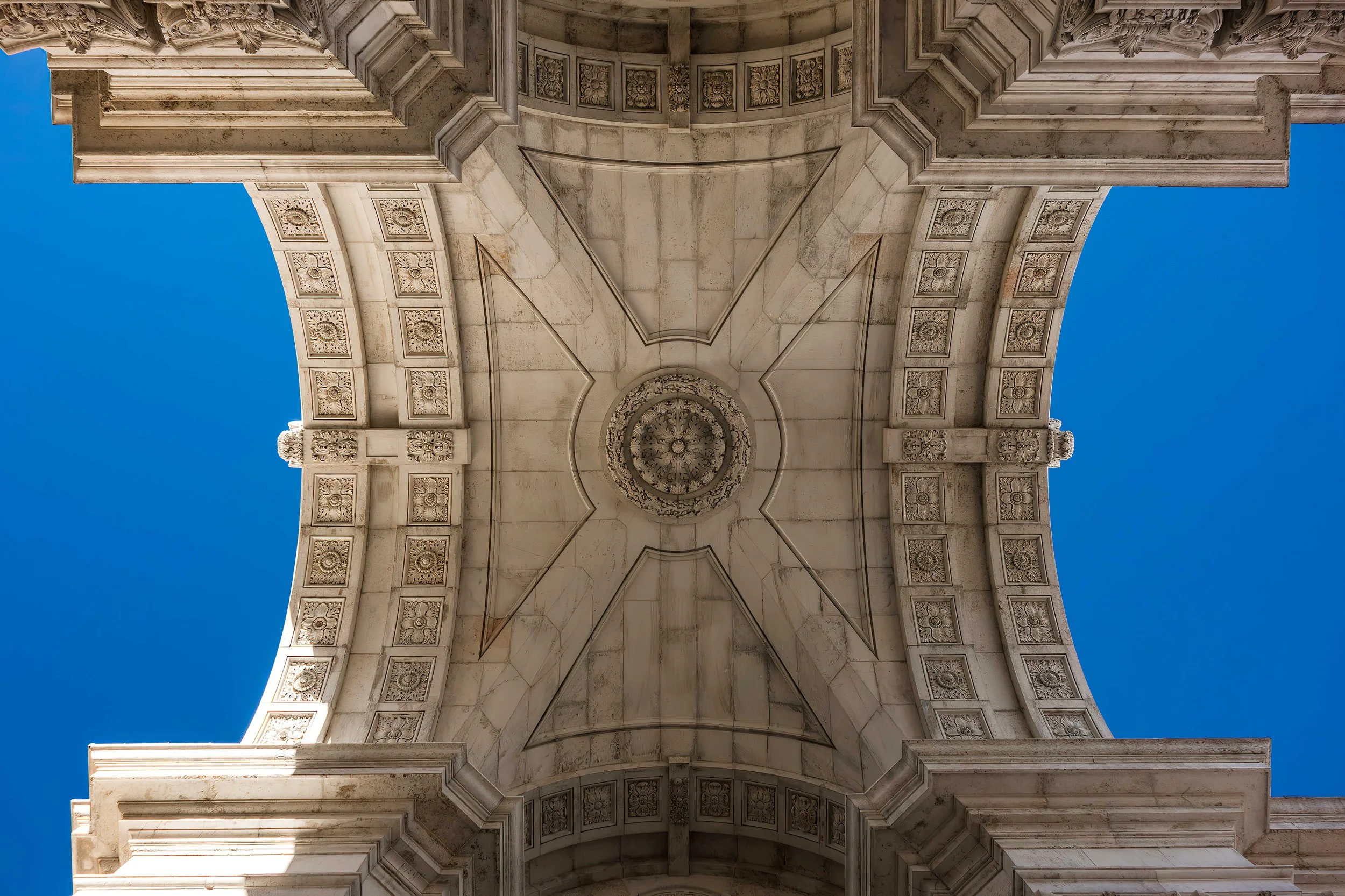 Looking up at the ornate ceiling of a stone building with detailed carvings and a central decorative medallion, against a clear blue sky.