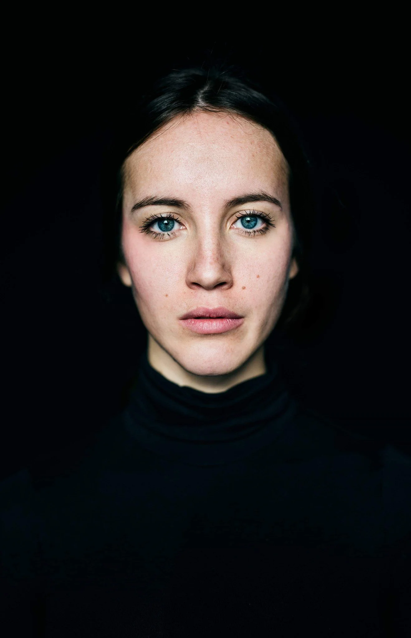 Close-up portrait of a young woman with bright blue eyes and dark hair, wearing a black turtleneck, against a solid black background.
