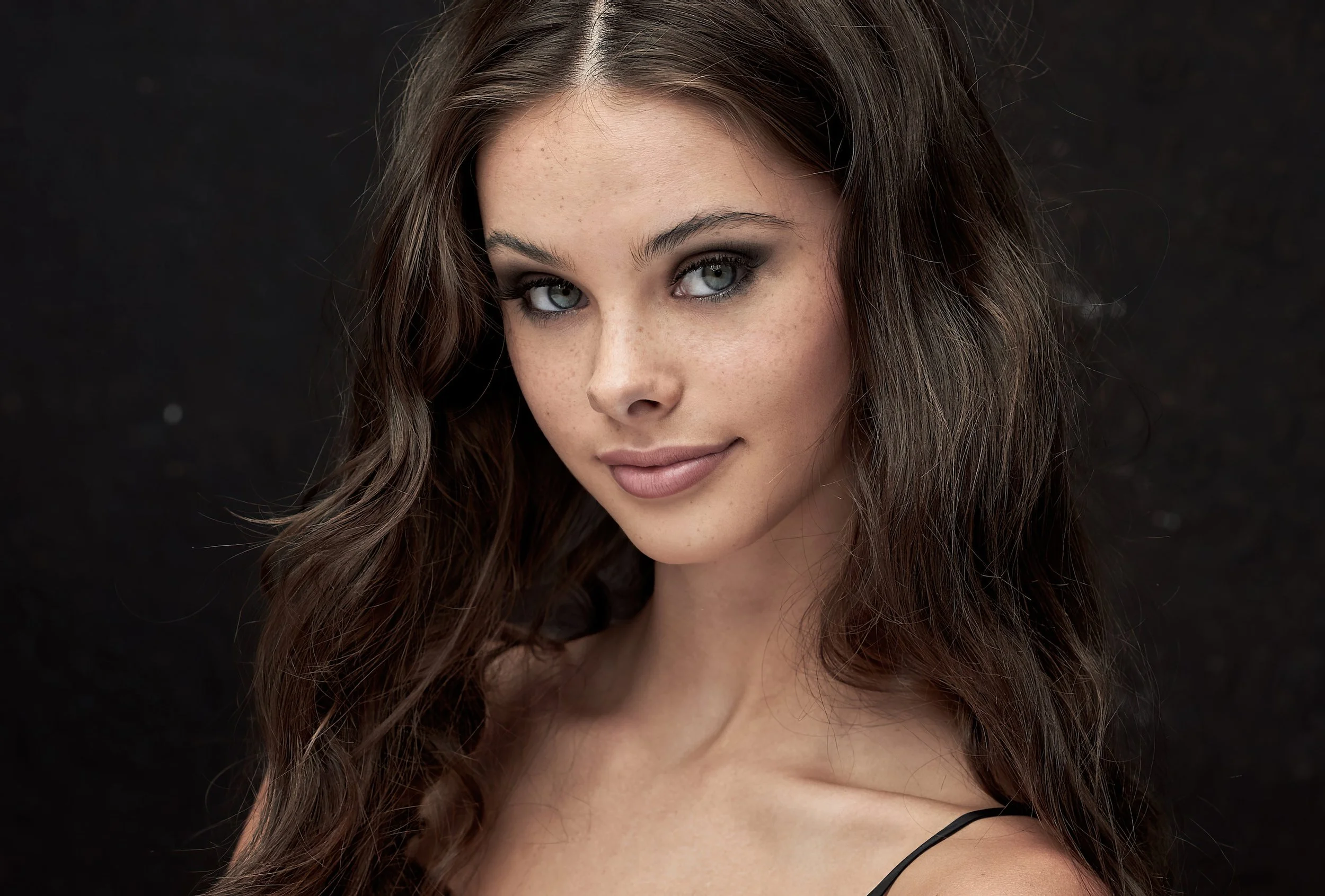 A young woman with long wavy brown hair, light skin, and blue eyes looking at the camera with a slight smile, wearing a black strap top, against a dark background.