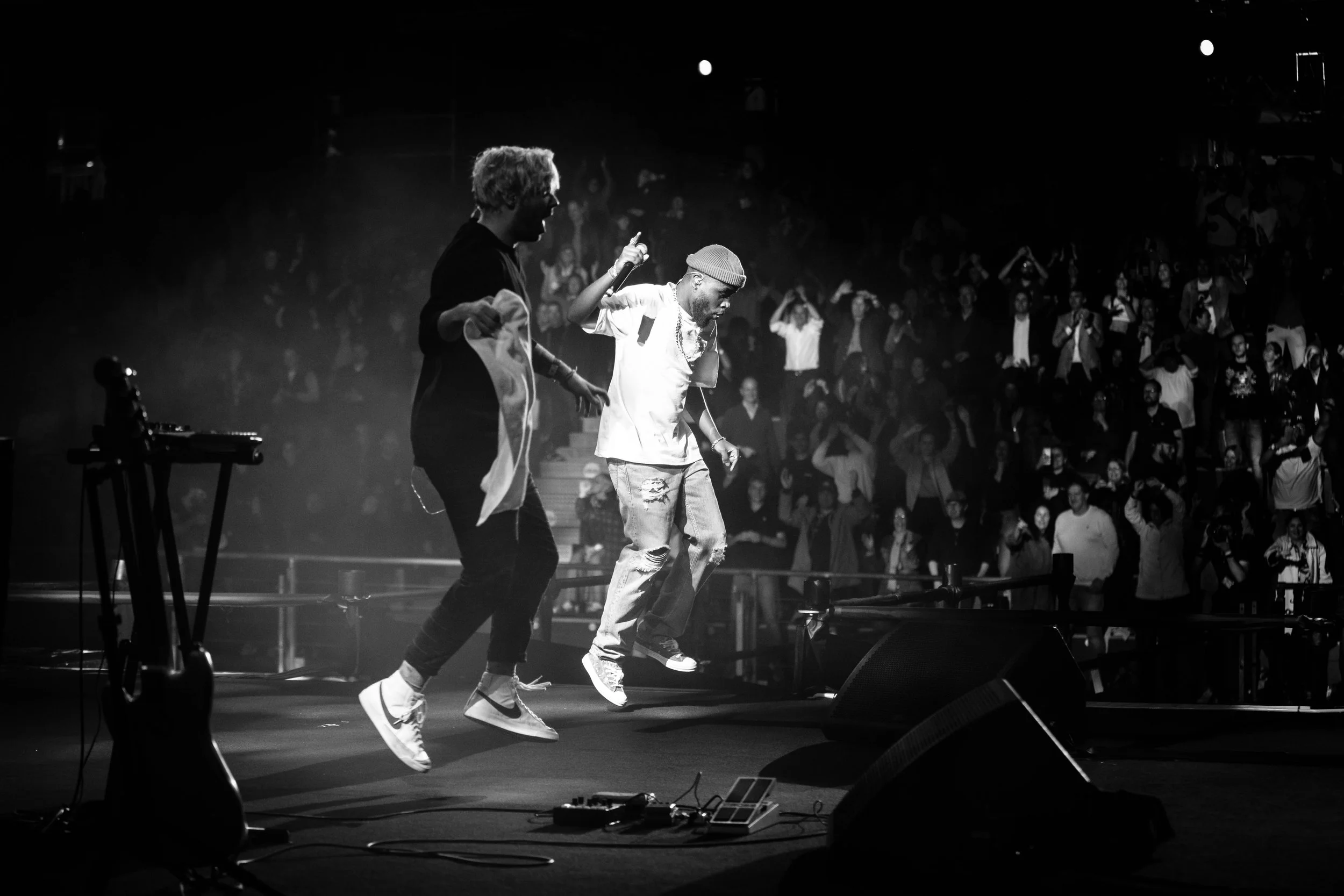 Two musicians dancing or jumping on a stage with a crowd in the background, black and white photo.